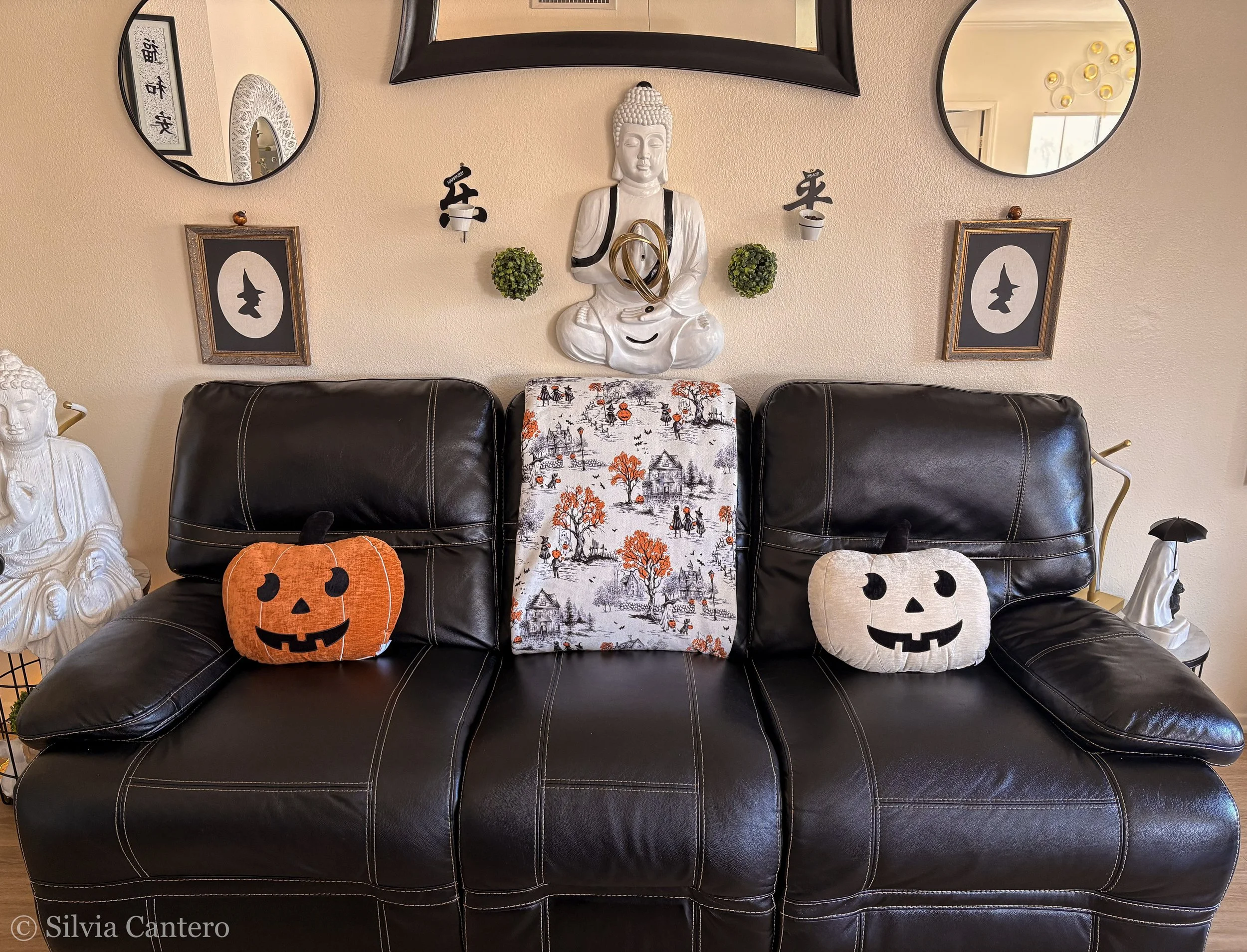 Decorative wall arrangement including a large white Buddha statue, two small green plants in white pots, framed pictures of witch silhouettes, hanging mirrors, and Halloween-themed plush pumpkins on a black leather sofa.