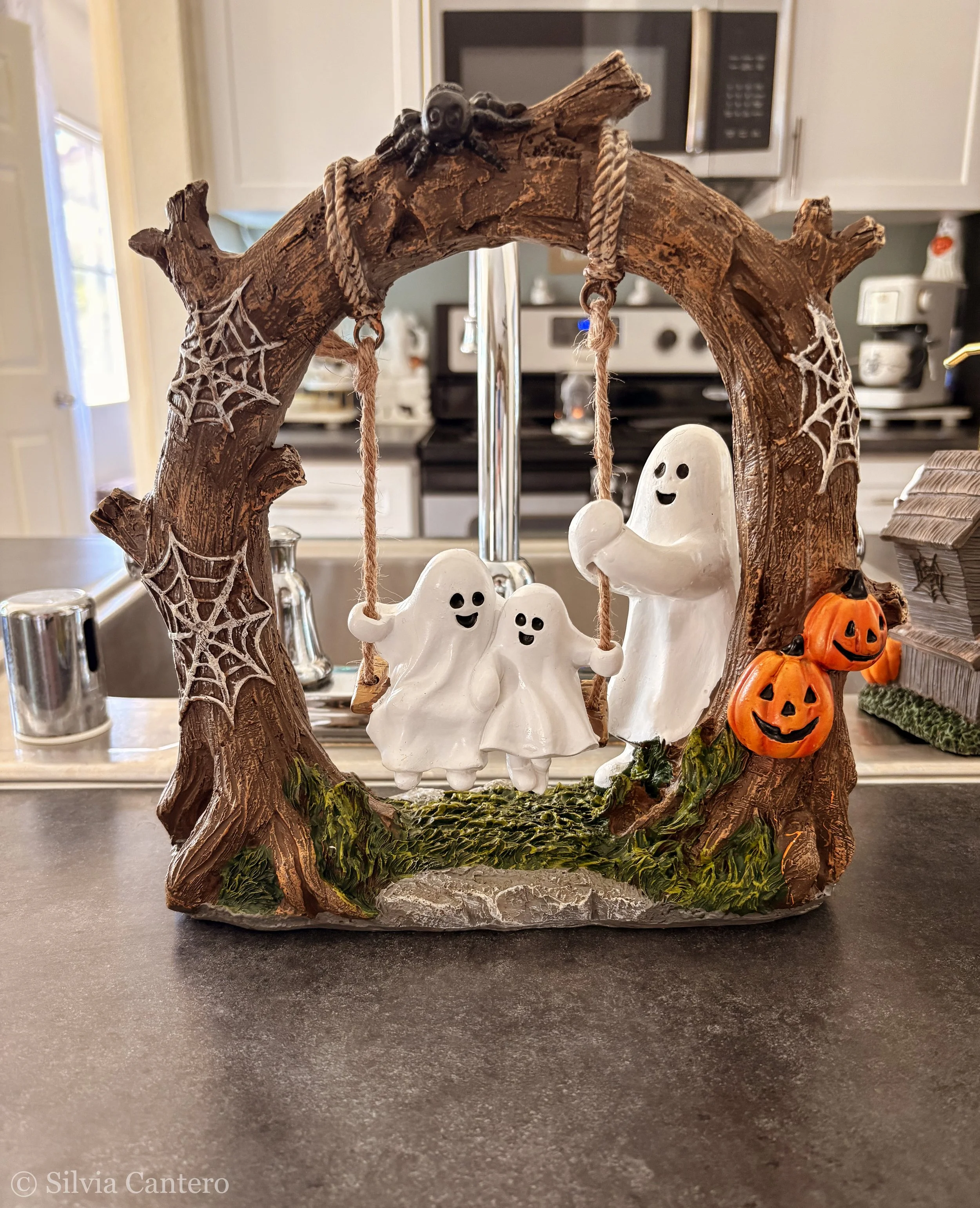 A Halloween-themed decorative ornament featuring a rustic wooden arch decorated with spider webs, hanging ghost ornaments with smiling faces, and small pumpkin decorations, set on a dark countertop in a kitchen.