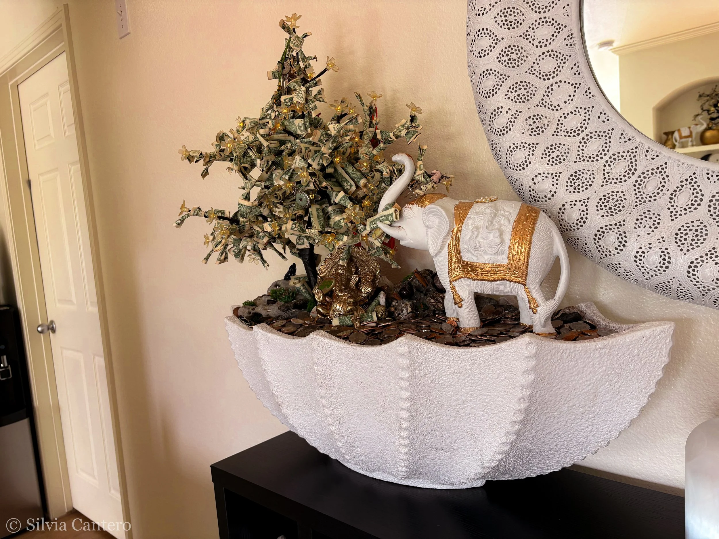 Decorative white elephant figurine with gold accents positioned next to a small artificial money tree made of rolled dollar bills and small gold figurines and flowers on a textured white table.