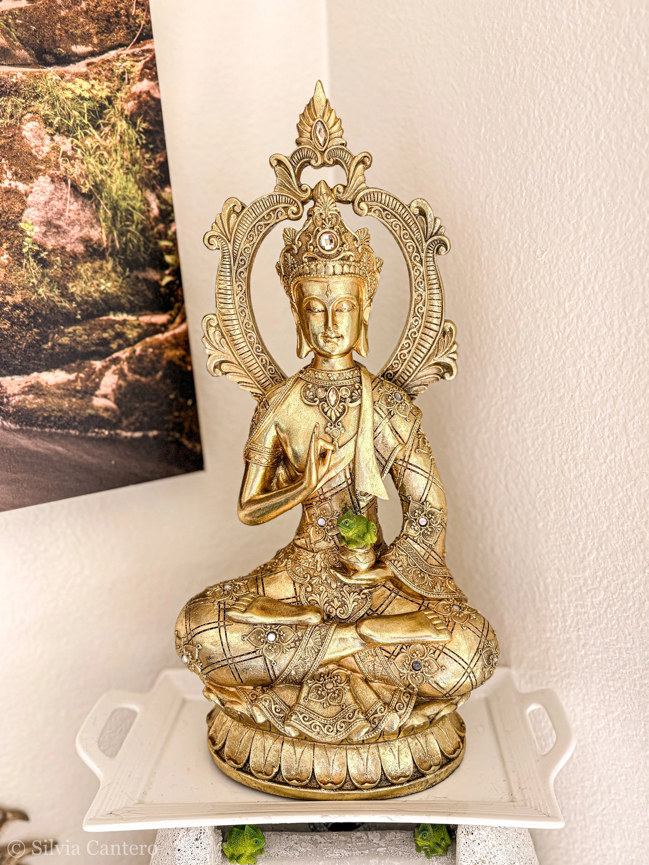 A golden statue of a deity with intricate details, seated in a meditative pose on a decorative base, displayed on a white platform against a plain wall with a partial photo of a nature scene on the left side.