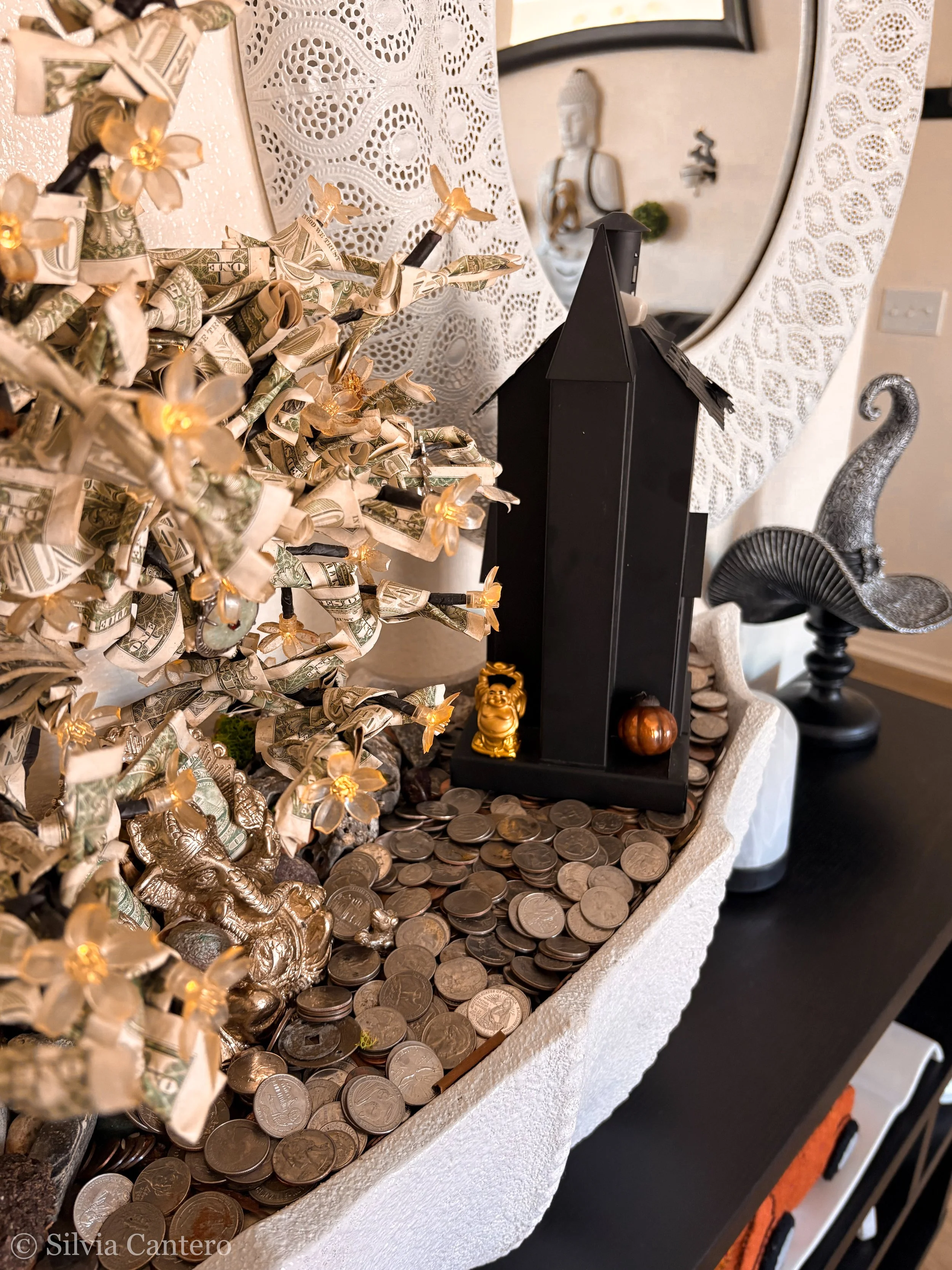 Decorative arrangement of folded dollar bills with small LED lights, positioned beside a mirror with Buddha and other statues, and a black structure with small gold and wood figurines, all on a surface scattered with coins.