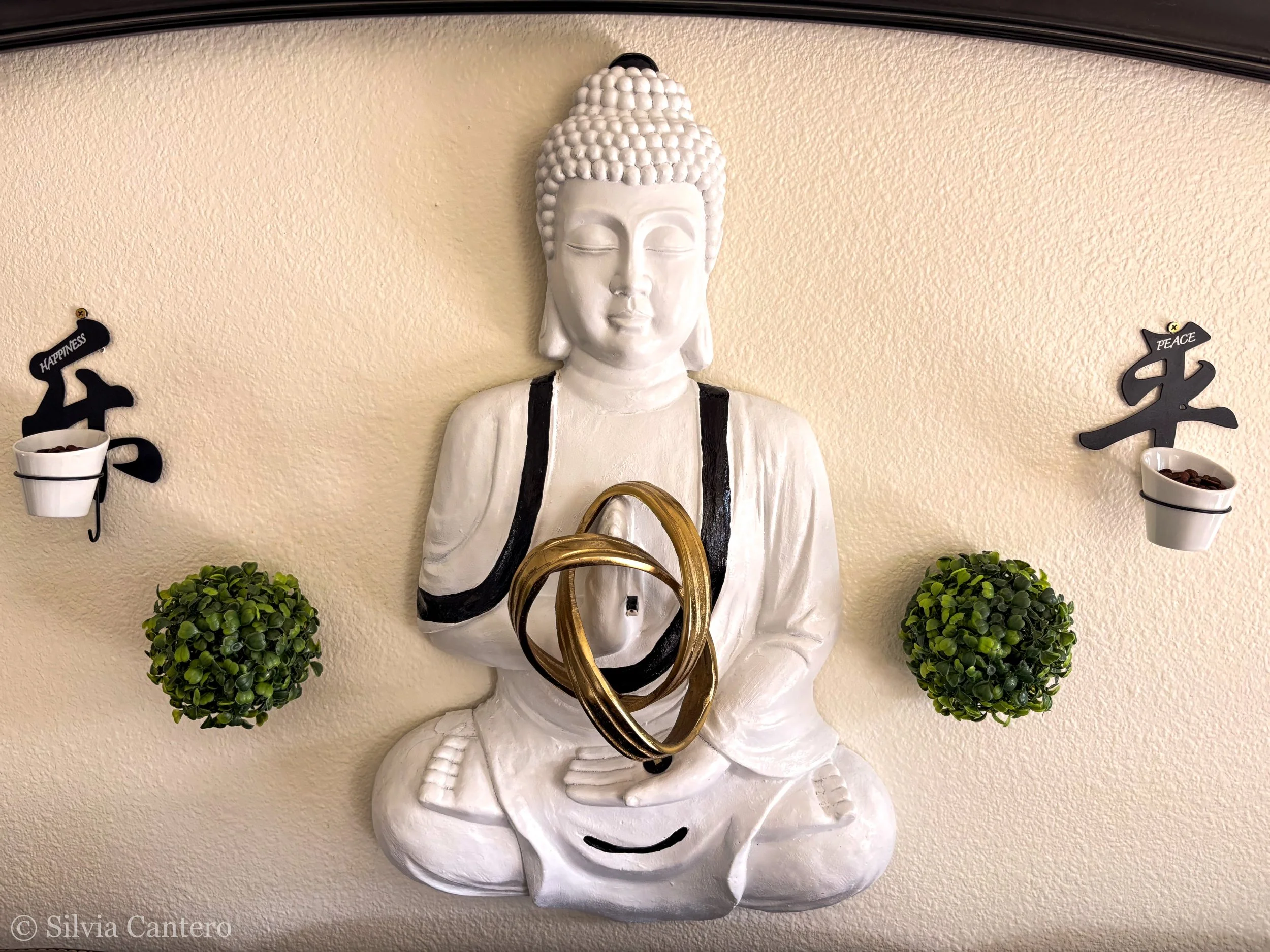 White Buddha statue with closed eyes holding a golden ring, flanked by two small green bushes and two white cups with black Japanese characters for 'happiness' and 'peace' on a beige wall.