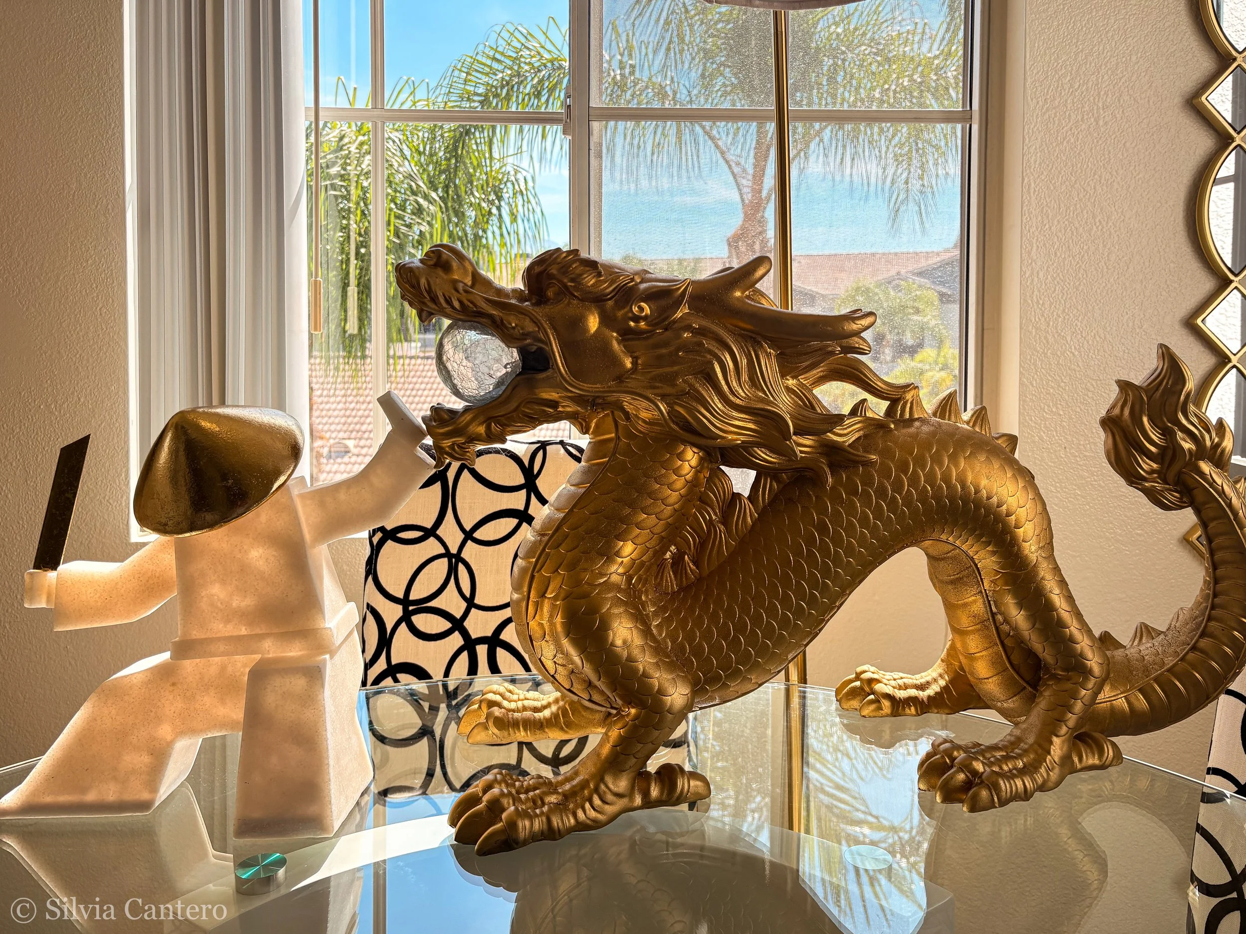 A golden dragon sculpture is displayed on a glass surface, behind a window showing palm trees and a blue sky. Part of a white and beige abstract sculpture is visible to the left.