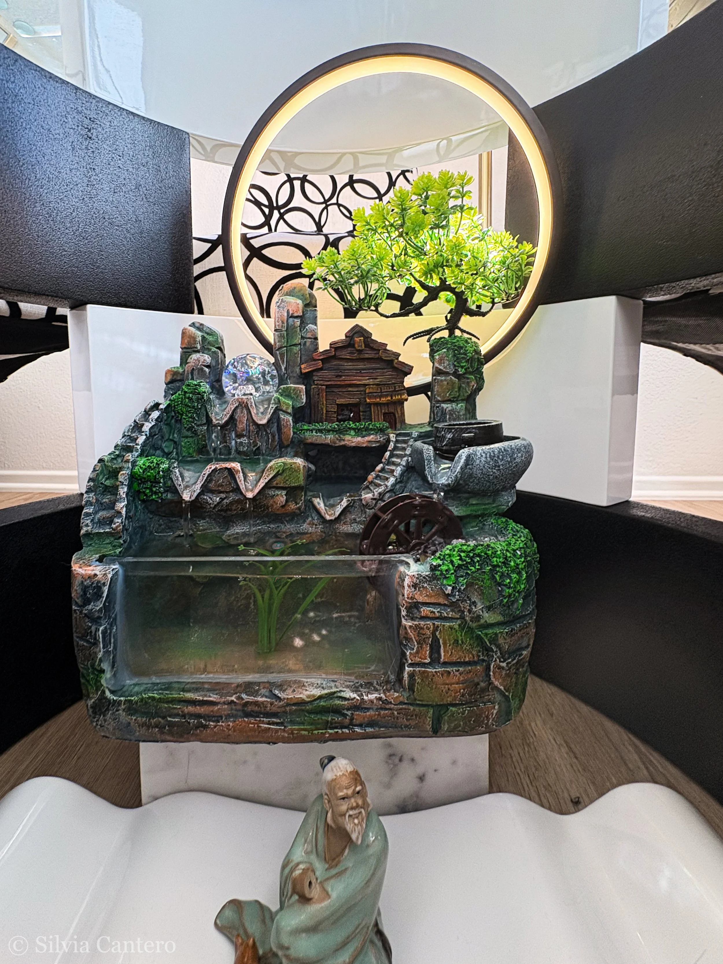 Decorative table with a balance of a lush green bonsai tree, a miniature wooden house, and a miniature pond with water and building miniatures, arranged around a circular LED mirror, with a black and white abstract patterned background and a figurine