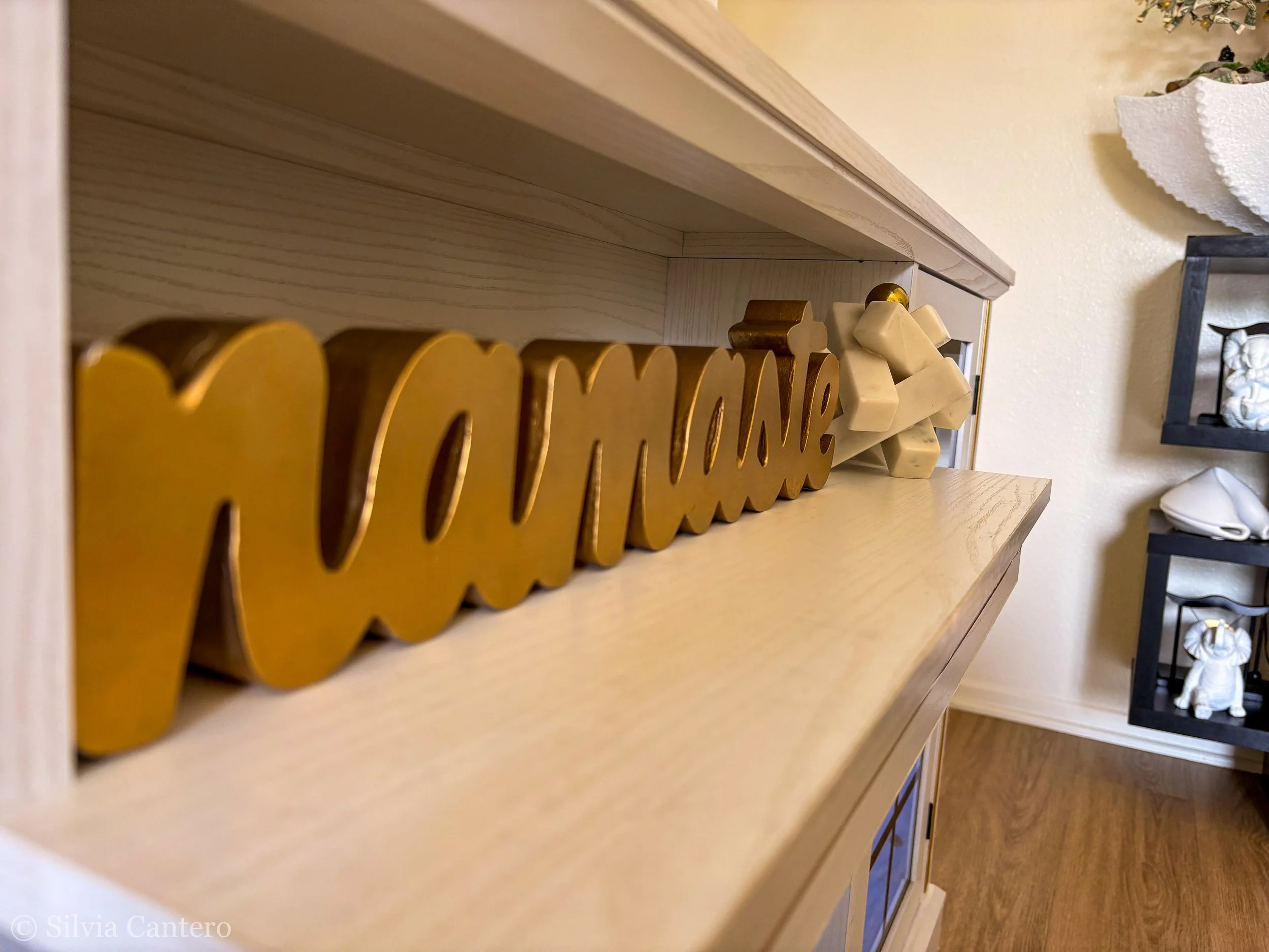 Close-up view of a wooden shelf with decorative items including a sign that says 'Love' in gold, some white geometric sculptures, and a small gold ball. Part of a black shelf with white decorative objects and a partial view of a room with beige walls