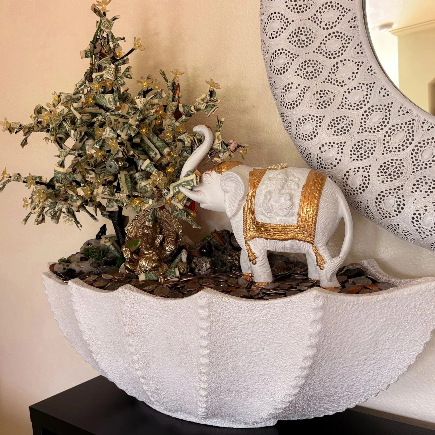 Decorative sculpture of a white elephant with gold accents, nose in a jar filled with coins, next to a money tree wrapped in dollar bills and gold ribbons, set on a white textured circular surface.