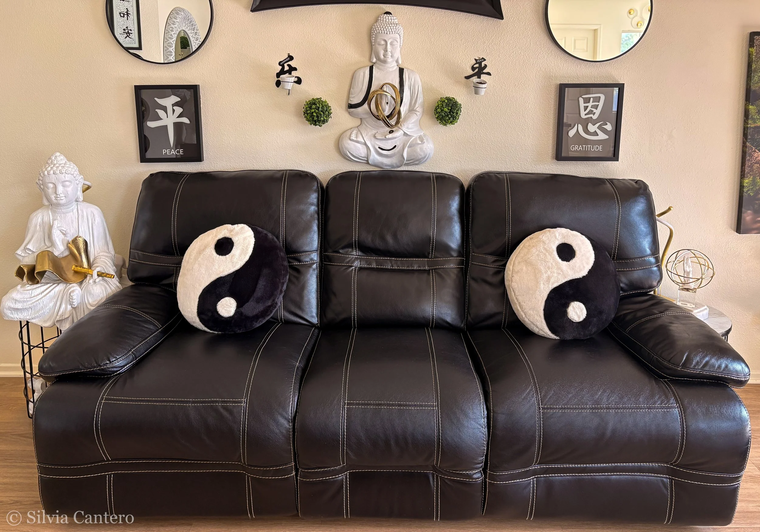 Living room wall decor featuring a large seated Buddha statue, framed peace and gratitude signs, two round mirrors, small plants, yin-yang pillows on a black leather sofa, and other decorative items.