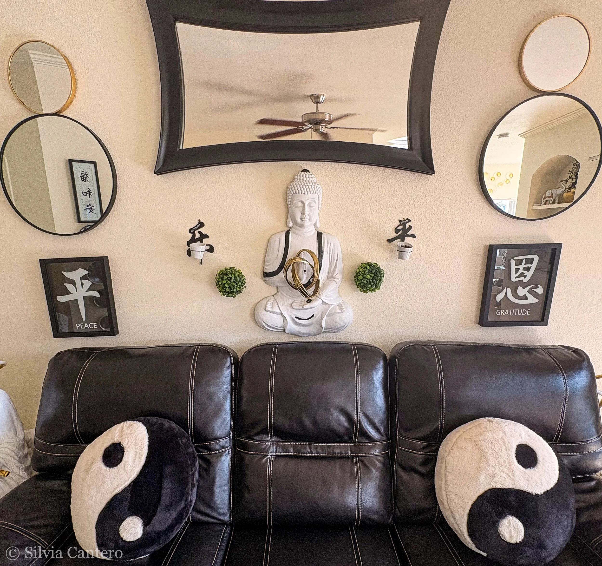 Living room wall decorated with four mirrors, a Buddha statue, framed art and plants on a black leather sofa.