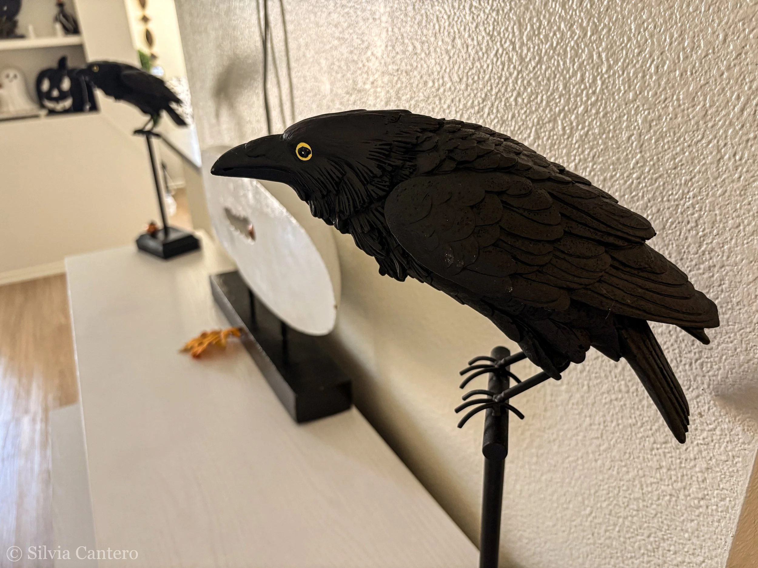 Black crow figurine on a stand in the foreground, with another similar crow figurine in the background on a shelf, set against a textured beige wall.