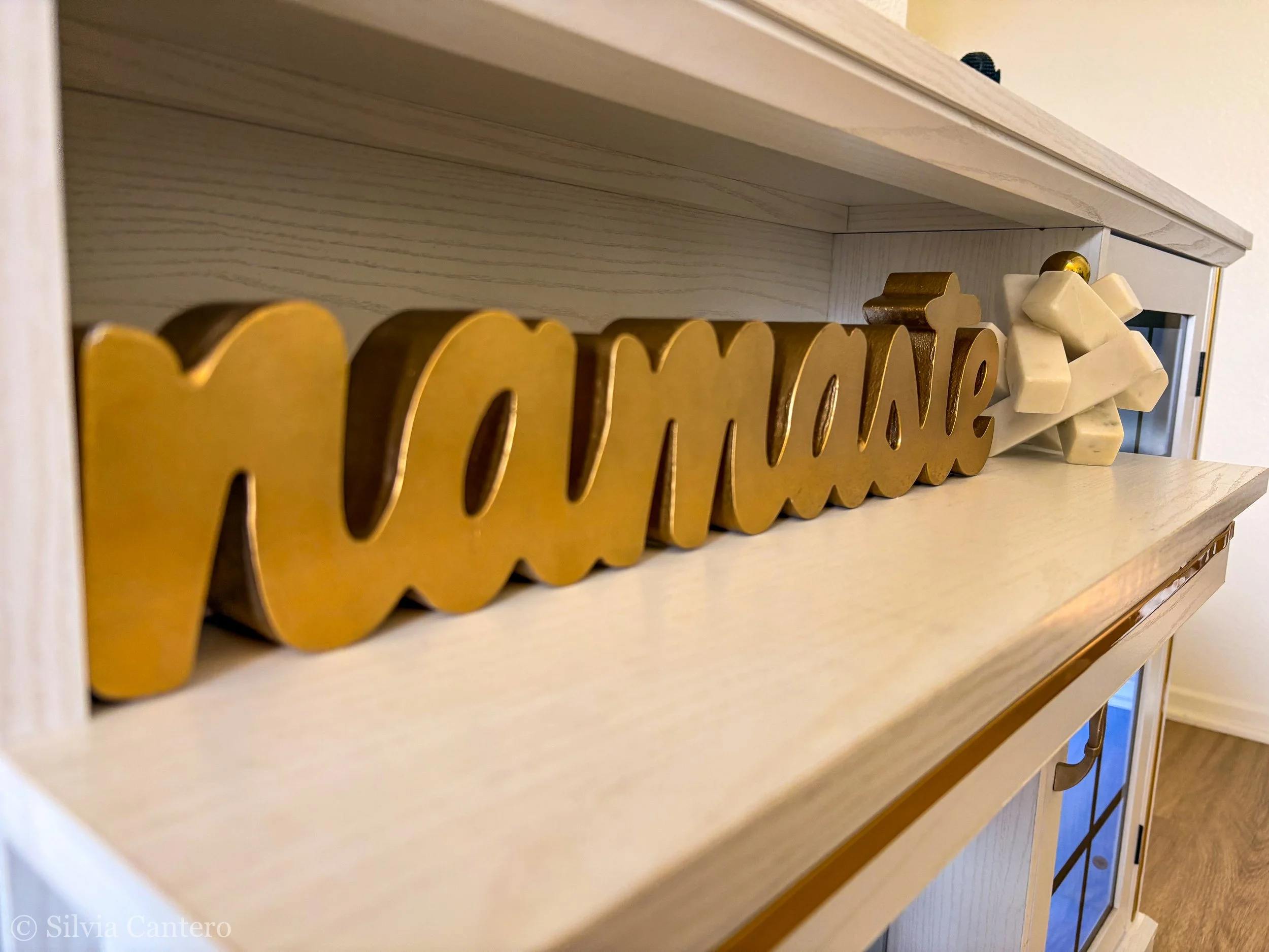 Wooden shelf with decorative blocks and a partial view of letters attached to the front, possibly spelling a word, with a cream wall in the background.