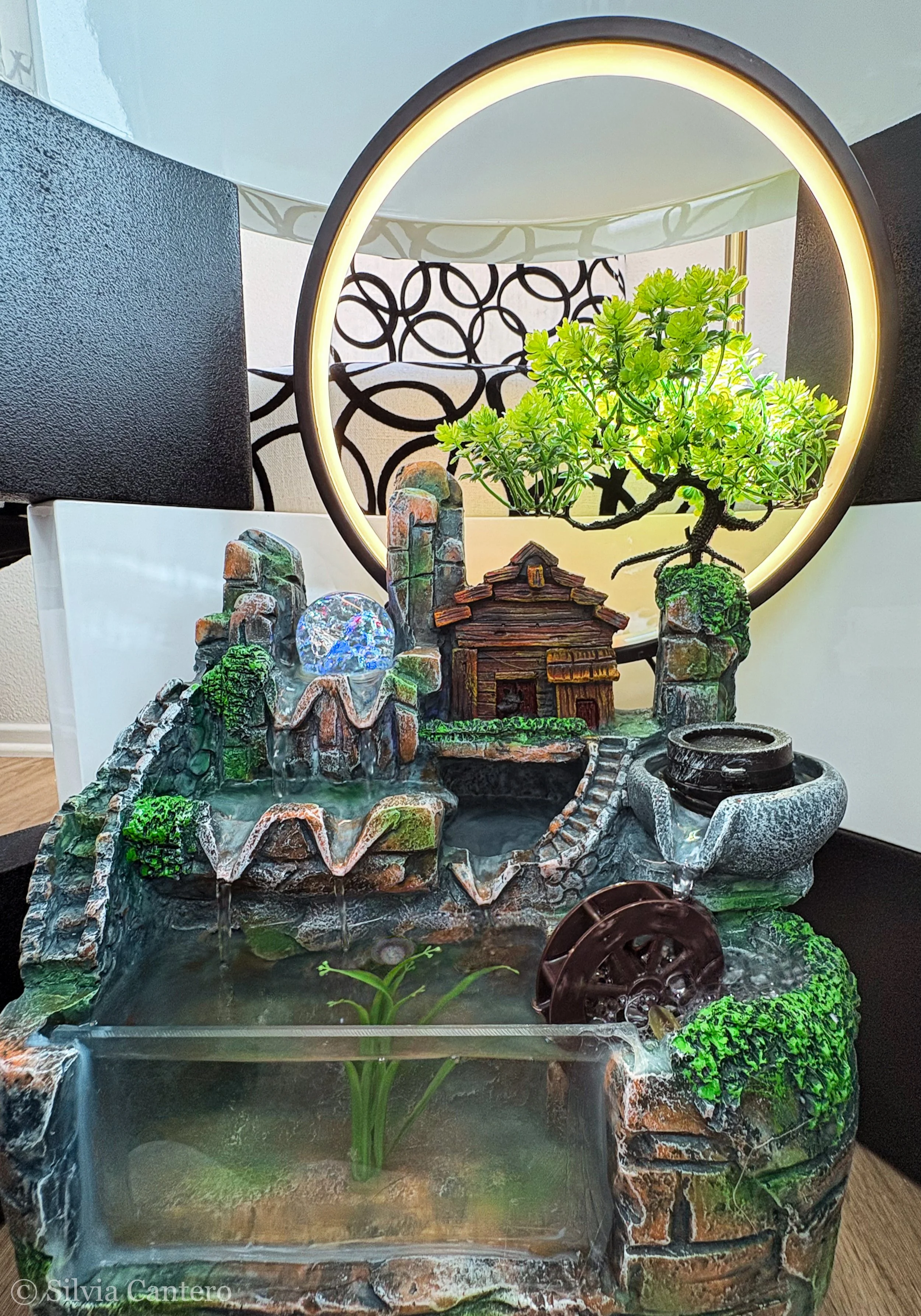 Miniature waterfall and pond with greenery, a small wooden house, and decorative rocks, illuminated by a ring light.