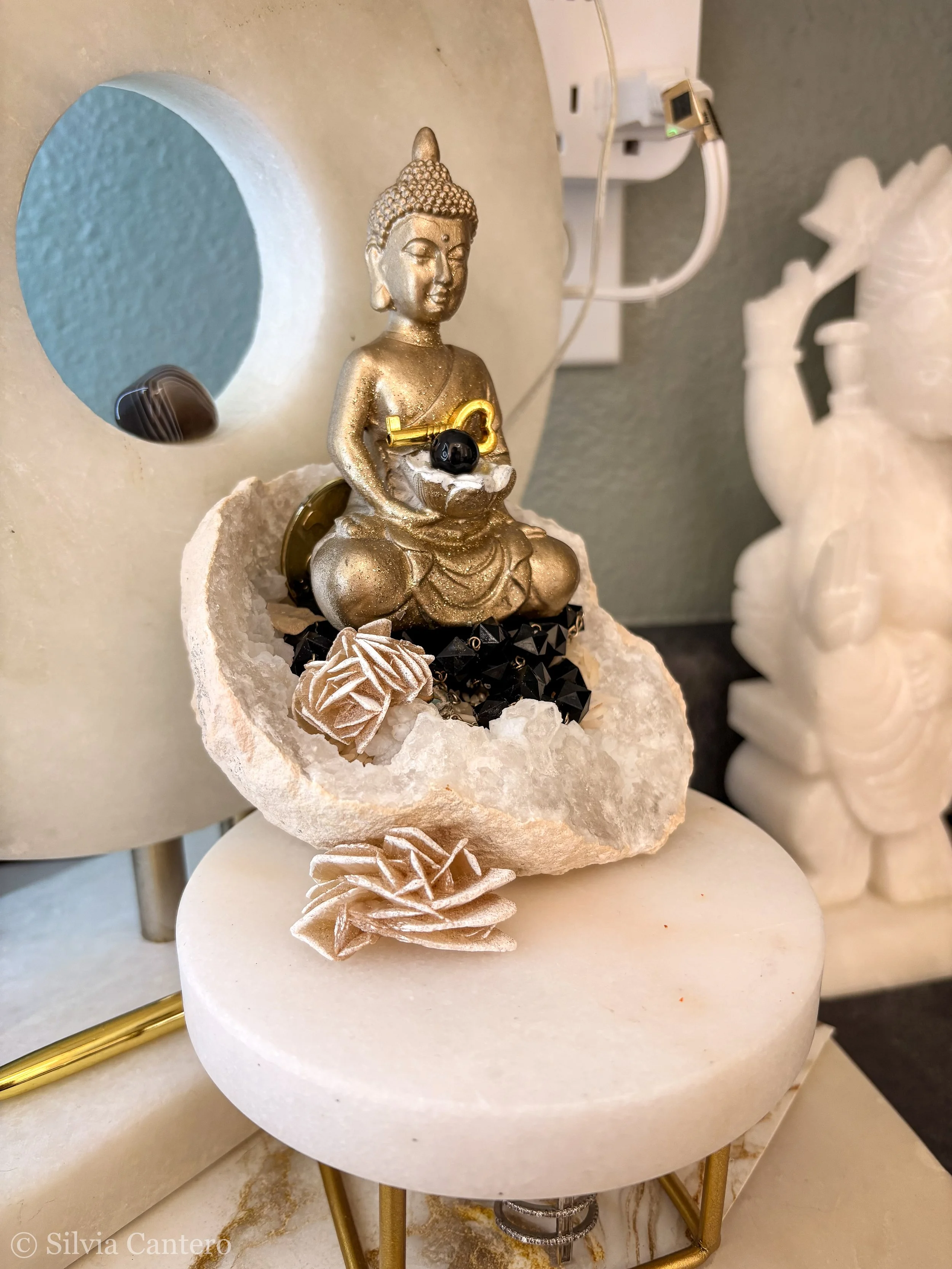 A decorative statue of Buddha sitting on a white stone base, holding a key and a black shiny stone, surrounded by small flowers and black beads. The statue is gold-colored with silver accents, and is positioned against a light-colored wall with elect