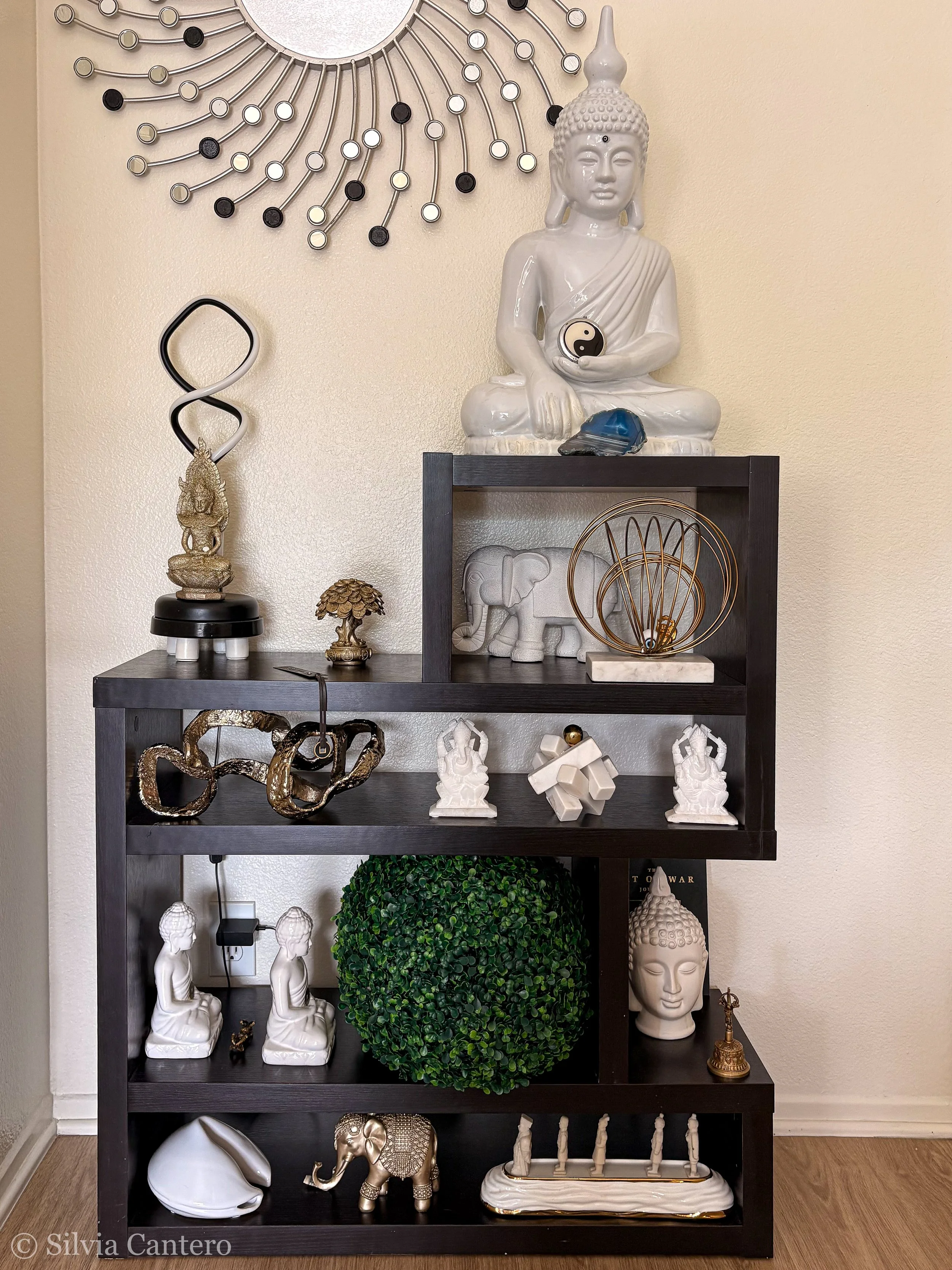 A black bookshelf with various decorative objects including Buddha statues, a small elephant sculpture, a golden decorative orb, and a plant. A large white Buddha head sculpture is on top of the bookshelf, and a Yin-Yang symbol is held by a reclining