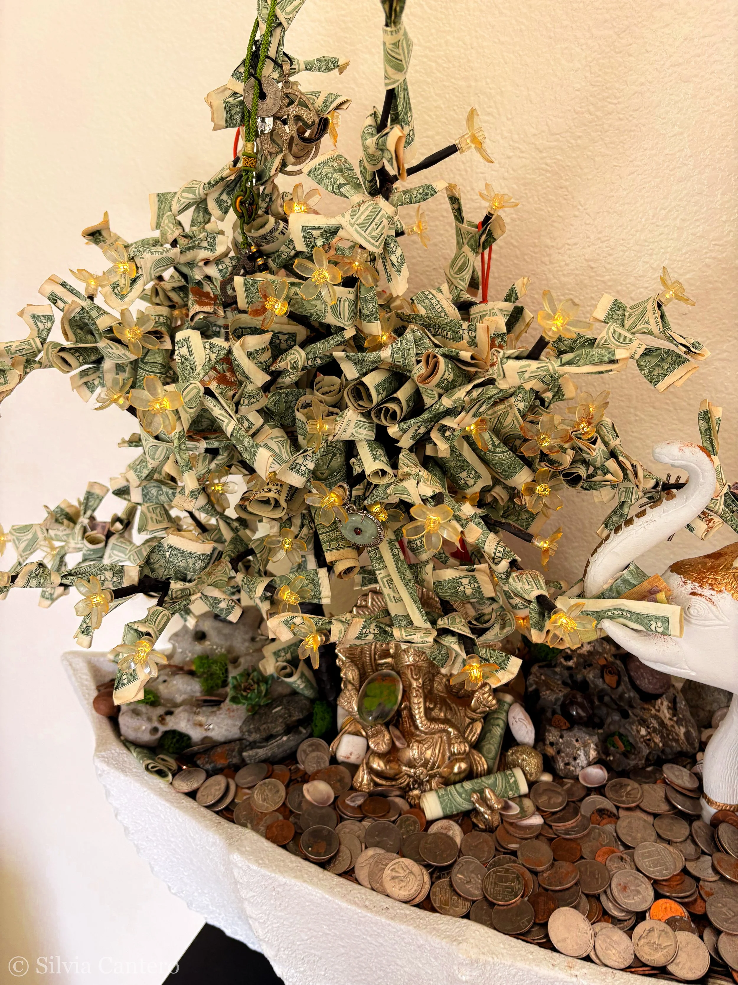 A decorative sculpture of a dragon covered with rolled dollar bills, surrounded by small fairy lights, coins, and figurines, on a white textured surface against a neutral-colored wall.