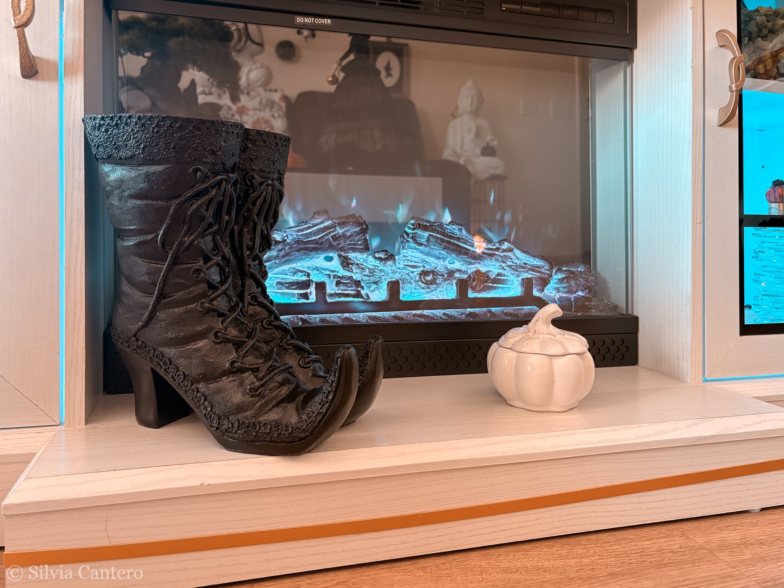 A black textured boot placed in front of a fireplace with blue flames and artificial logs. The fireplace has a glass cover reflecting two people sitting across from it, a woman in white attire and a person with a hat. To the right, a white decorative