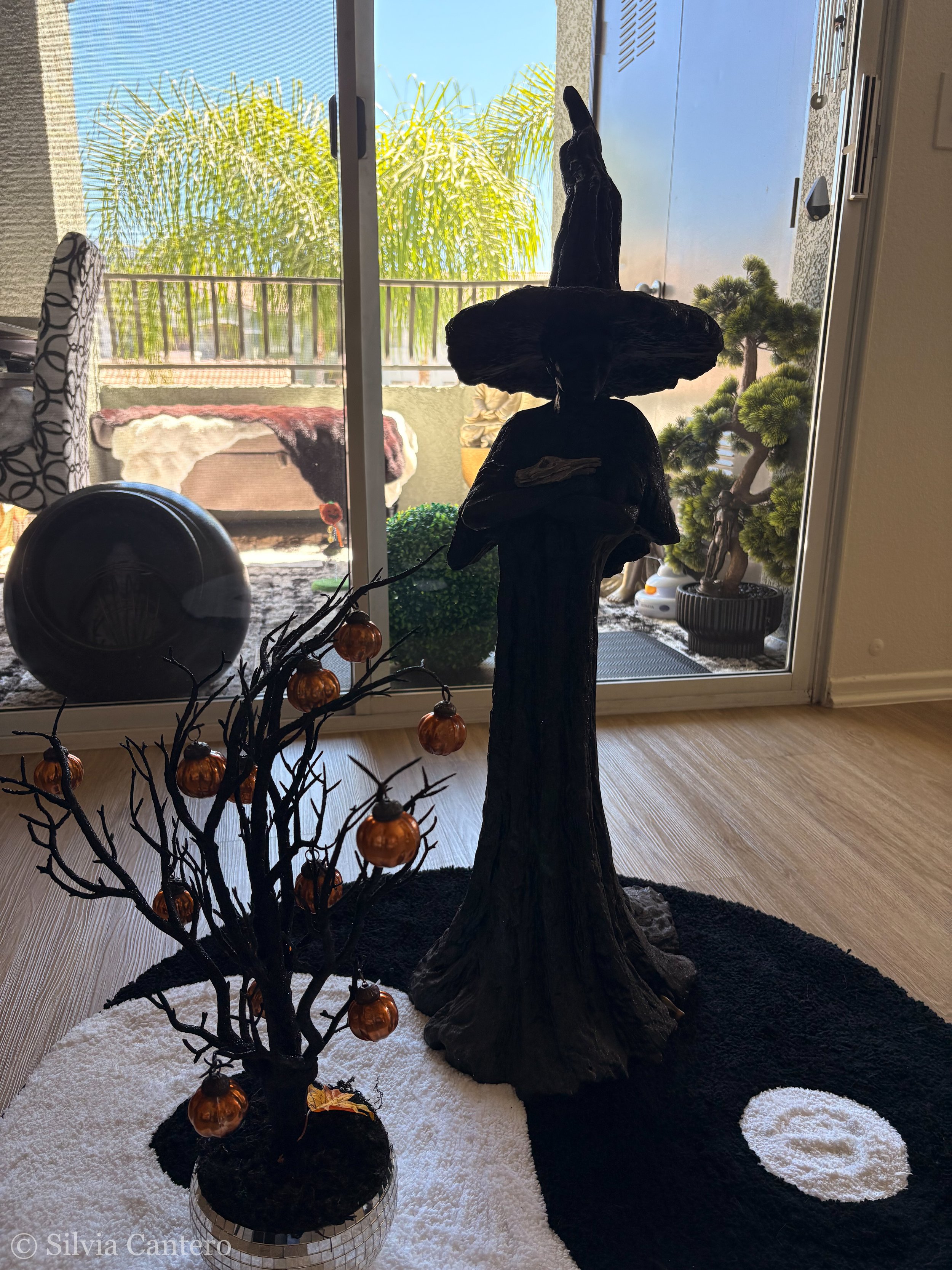 A decorative indoor scene with a black angel statue, a small decorative tree with orange ornaments, a black and white circular rug, and a sliding glass door leading to a balcony with a view of green trees and a blue sky.
