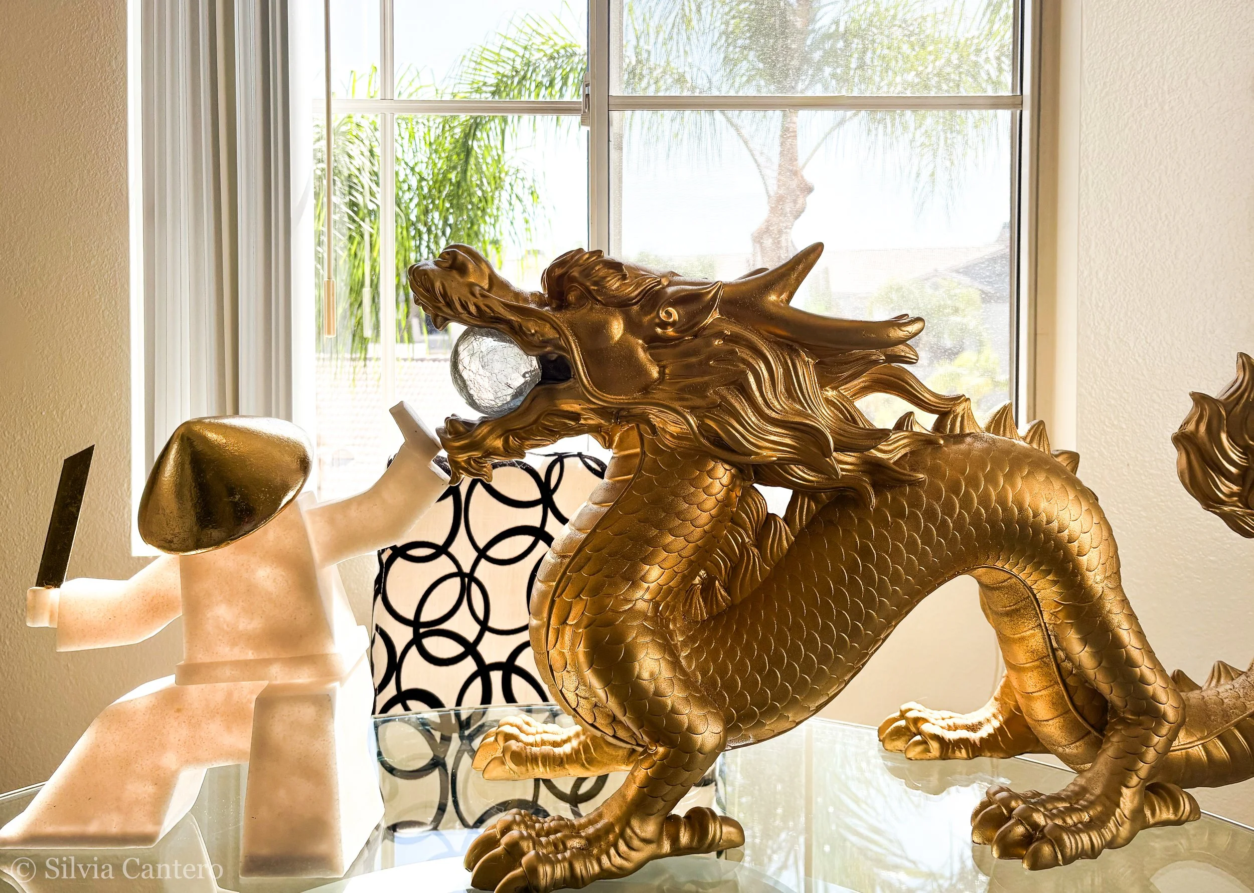 Gold-colored dragon sculpture with detailed scales, horns, and mane, positioned indoors near a window with palm trees outside, alongside a white marble bust, a decorative lattice, and a glass surface.