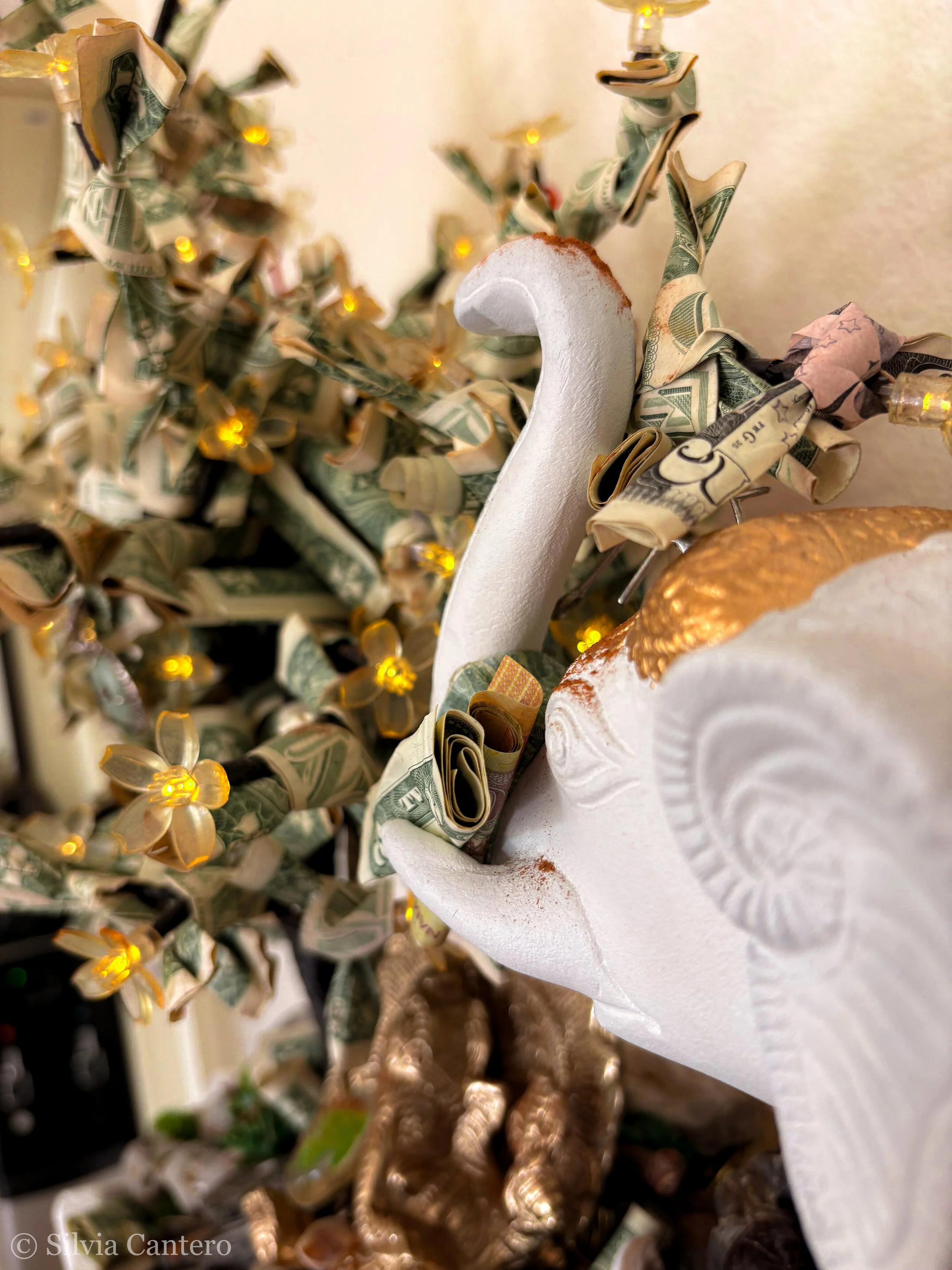 Close-up of a swan-shaped lamp base decorated with rolled dollar bills and small yellow lights, with a blurred background of a Christmas tree decorated with dollar bill ornaments and yellow lights.