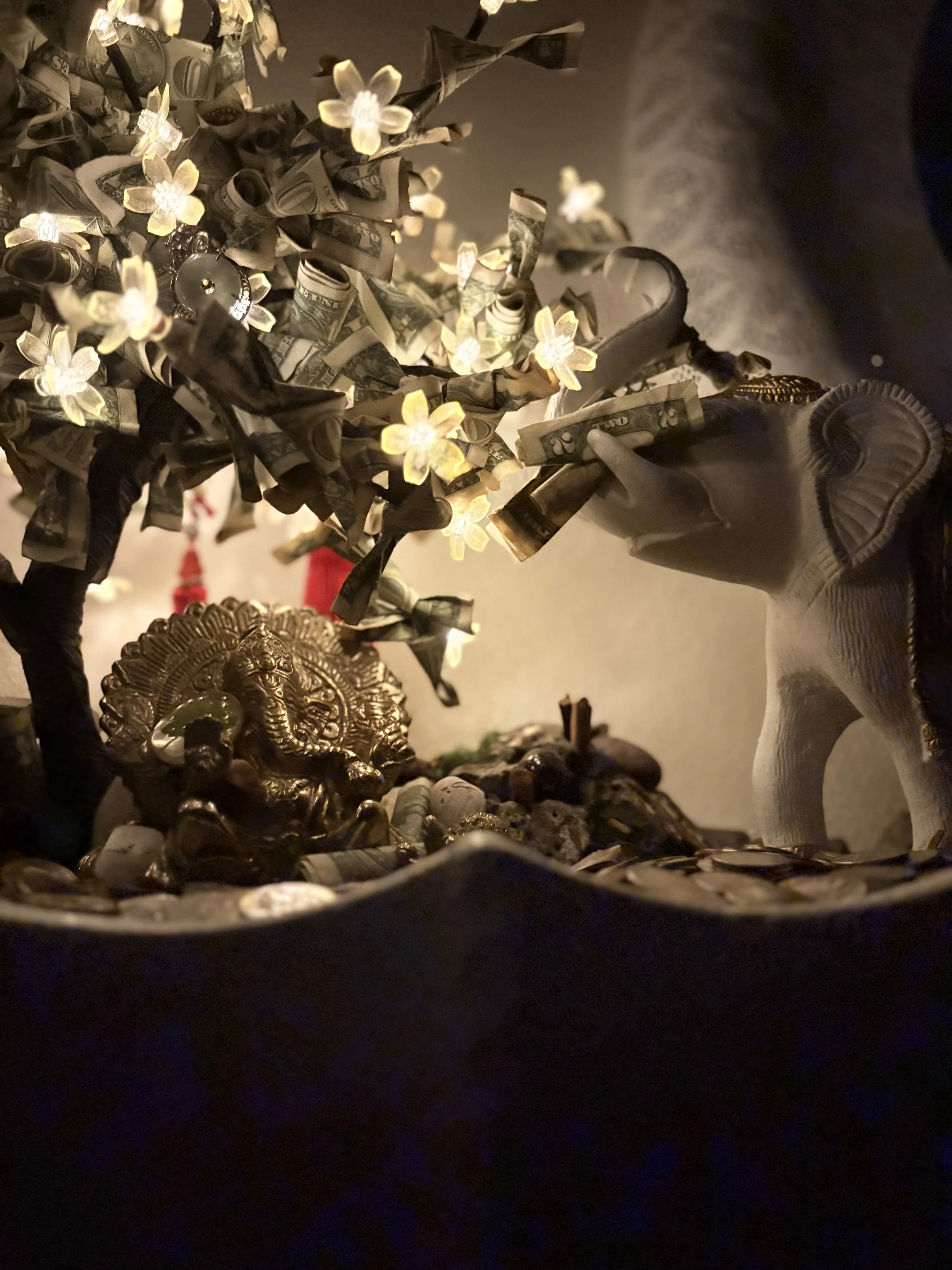 A small decorative tree wrapped in dollar bills and illuminated with small flower-shaped lights, with an ornate metallic lion figure in the foreground.