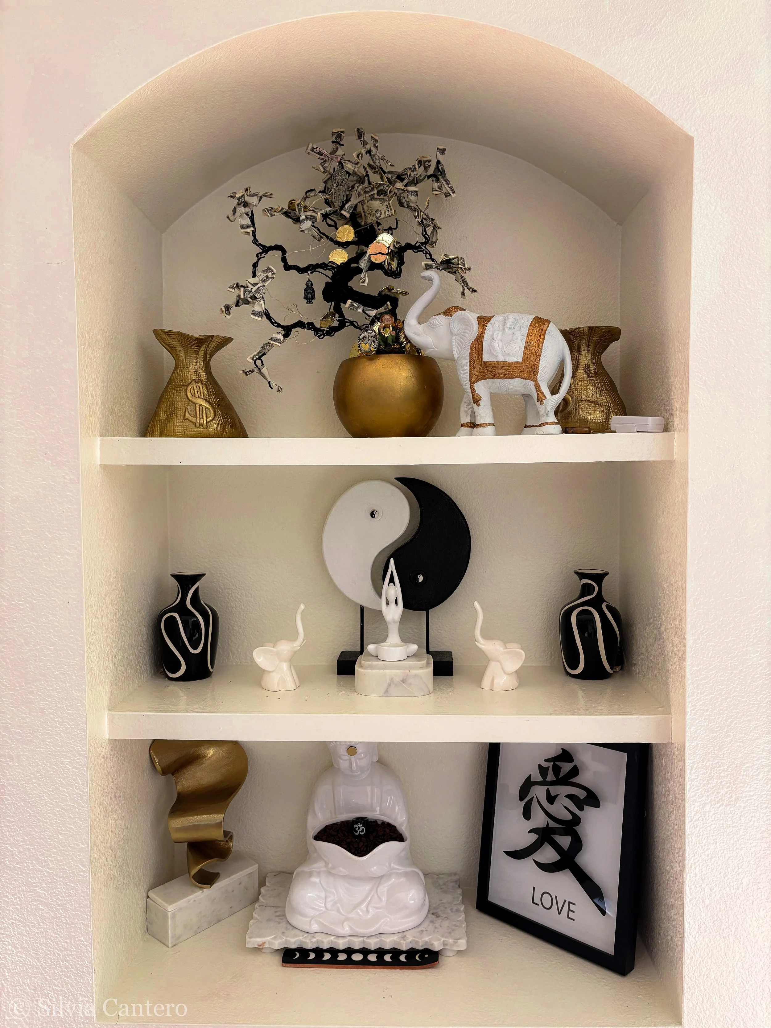 Decorative shelf with a money tree made of paper dollar bills in a gold pot, a white elephant figurine with a gold saddle, two gold vases with dollar signs, yin-yang symbol, small elephant and human figurines, a black and white framed love quote with