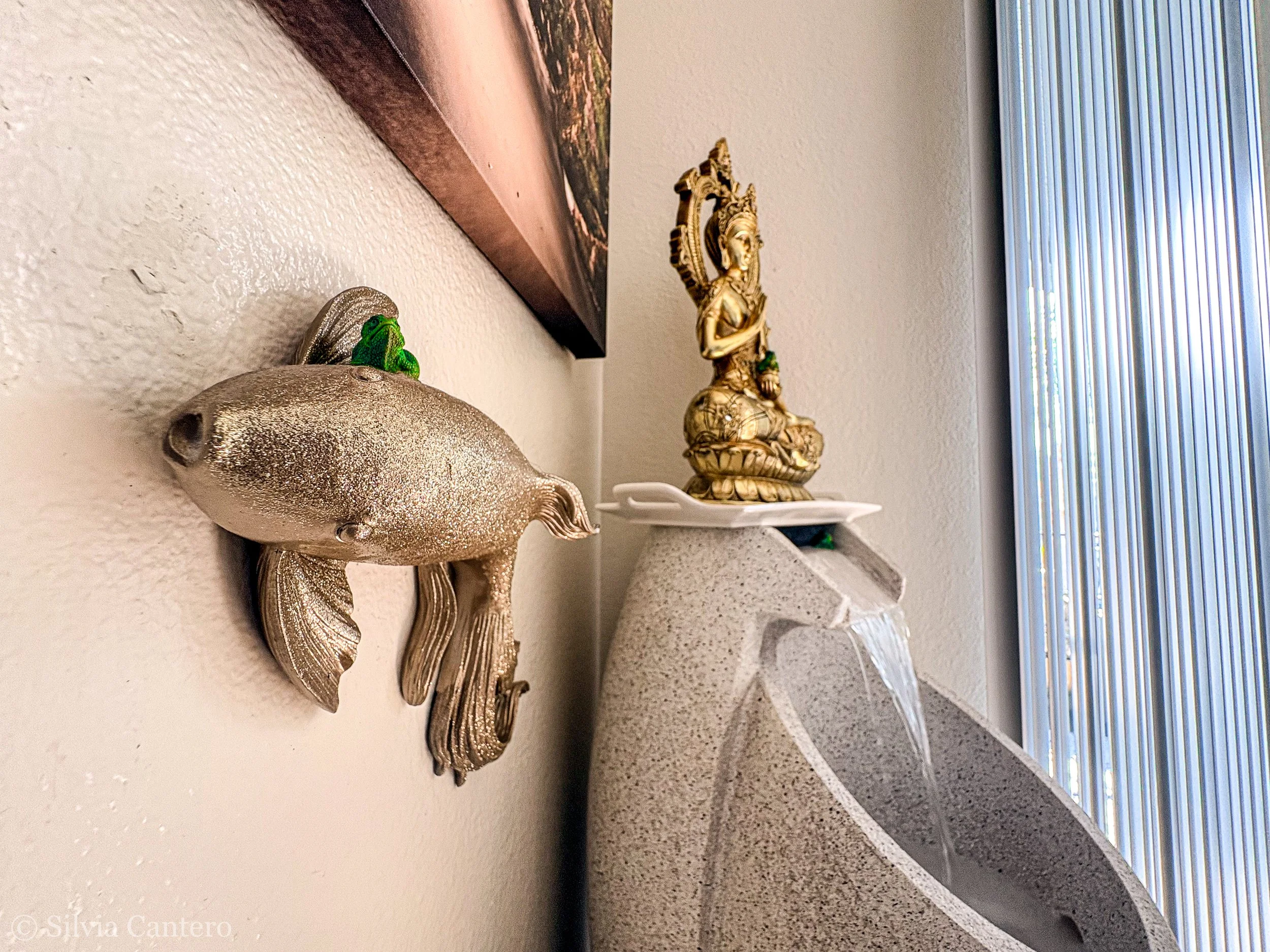 Decorative fish wall ornament, golden religious statue with multiple tiers, white plate beneath it, window with blinds in background.