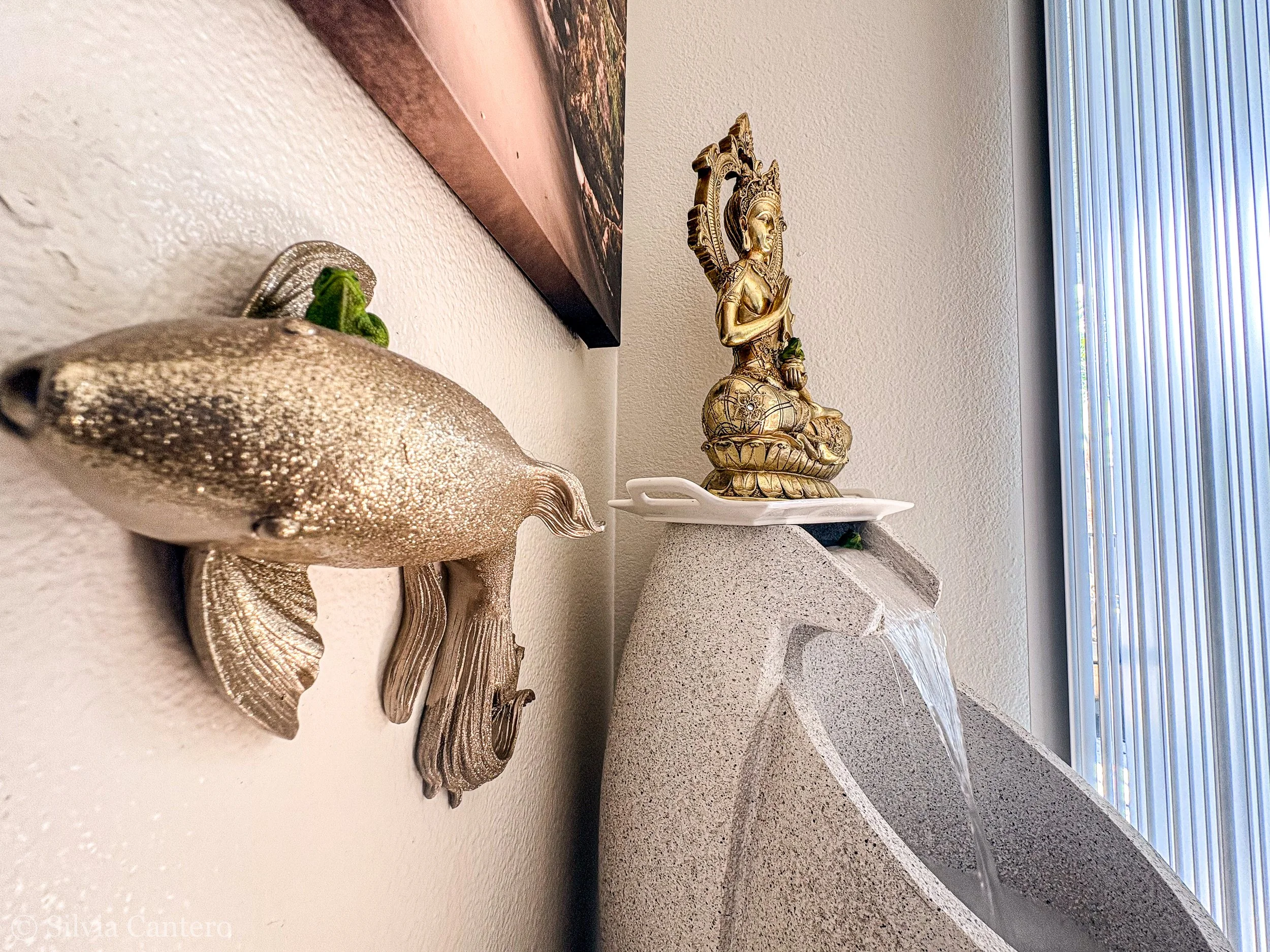 Decorative fish sculpture on a white shelf and a gold-colored figurine of a woman with an elaborate headdress, sitting on a carved platform, in front of a window with vertical blinds.