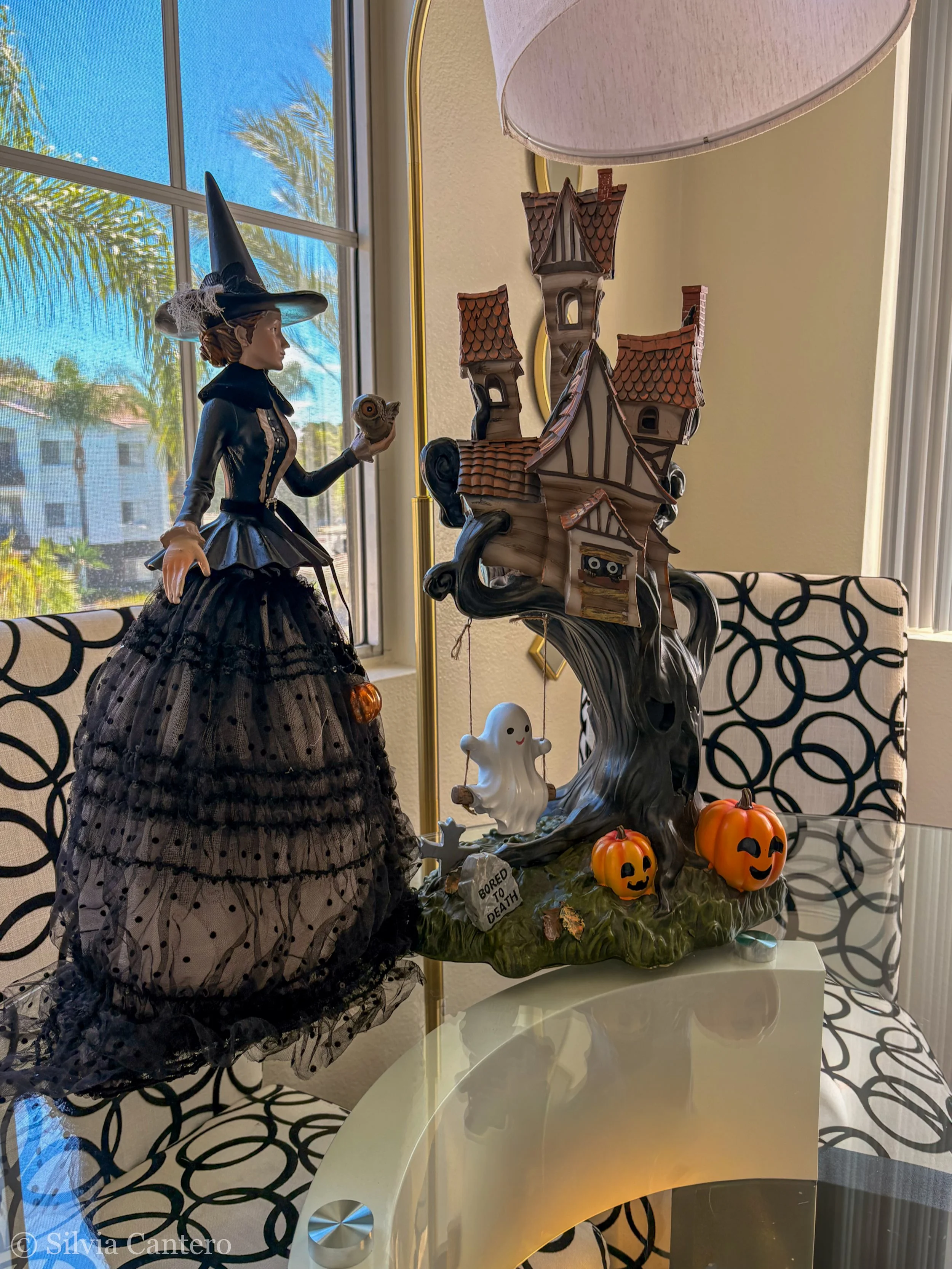 A Halloween-themed Witch figurine holding a scroll, beside a decorative haunted house with jack-o'-lanterns, a ghost, and a small sign that reads "BORED TO DEATH," on a white table near a window with palm trees outside.
