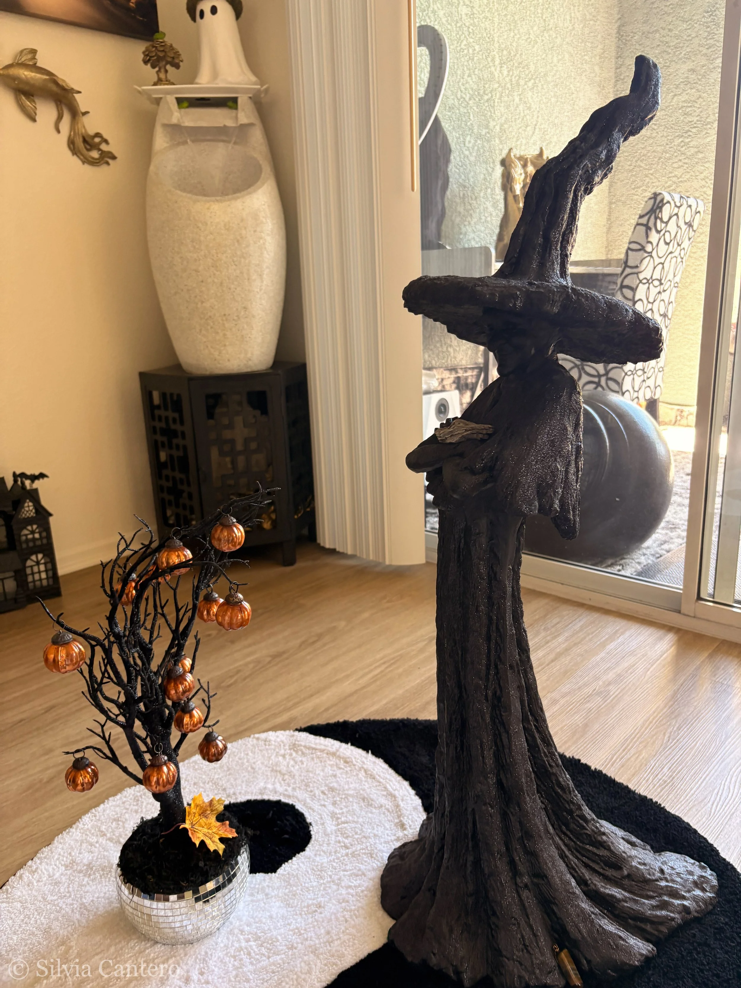 Indoor Halloween decoration scene featuring a large black fairy tale tree sculpture, a small black decorative tree with orange pumpkin lights, and a disco ball, set on a white circular rug with black accents. In the background, there are various home