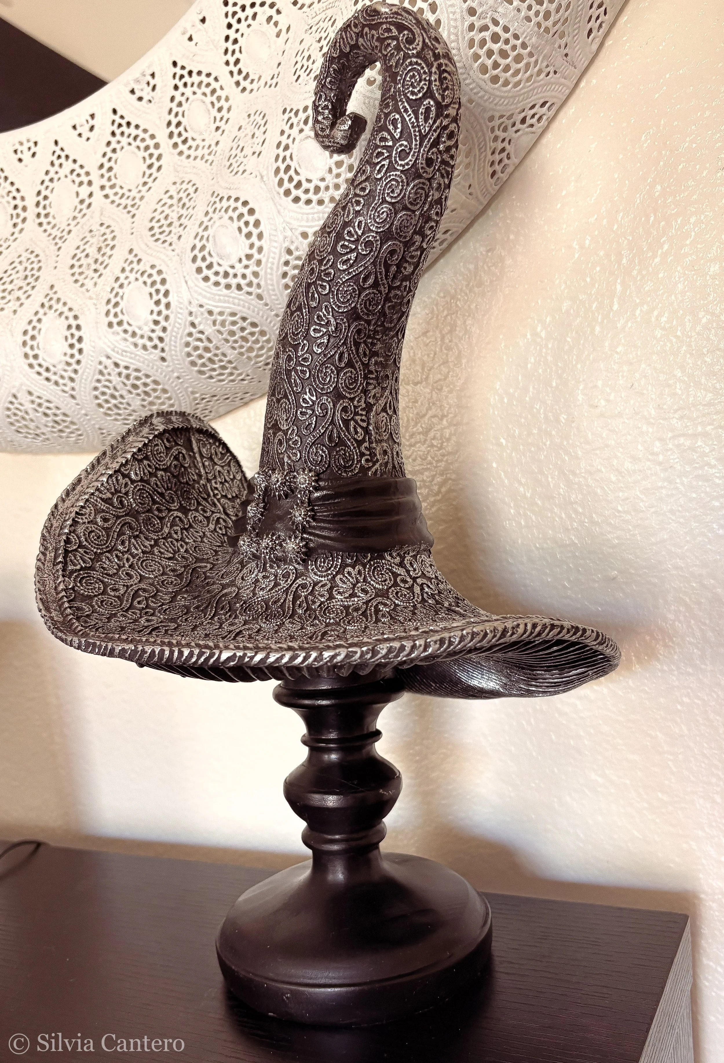 Decorative silver and black sculpture of an angel figure with detailed swirling patterns, placed on a dark wooden pedestal table.