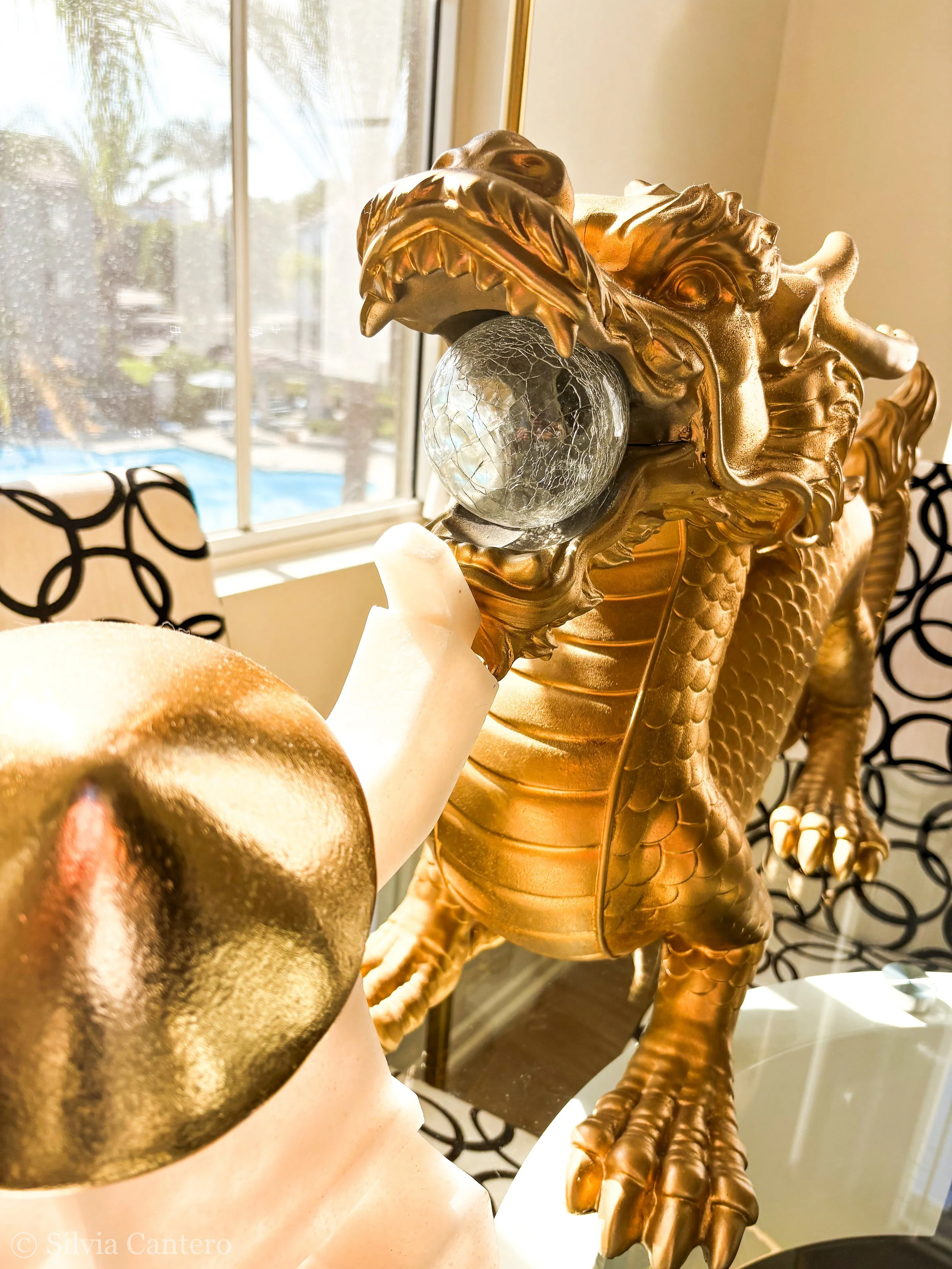 A golden dragon sculpture with a crystal ball in its mouth, set near a window with sunlight shining through.