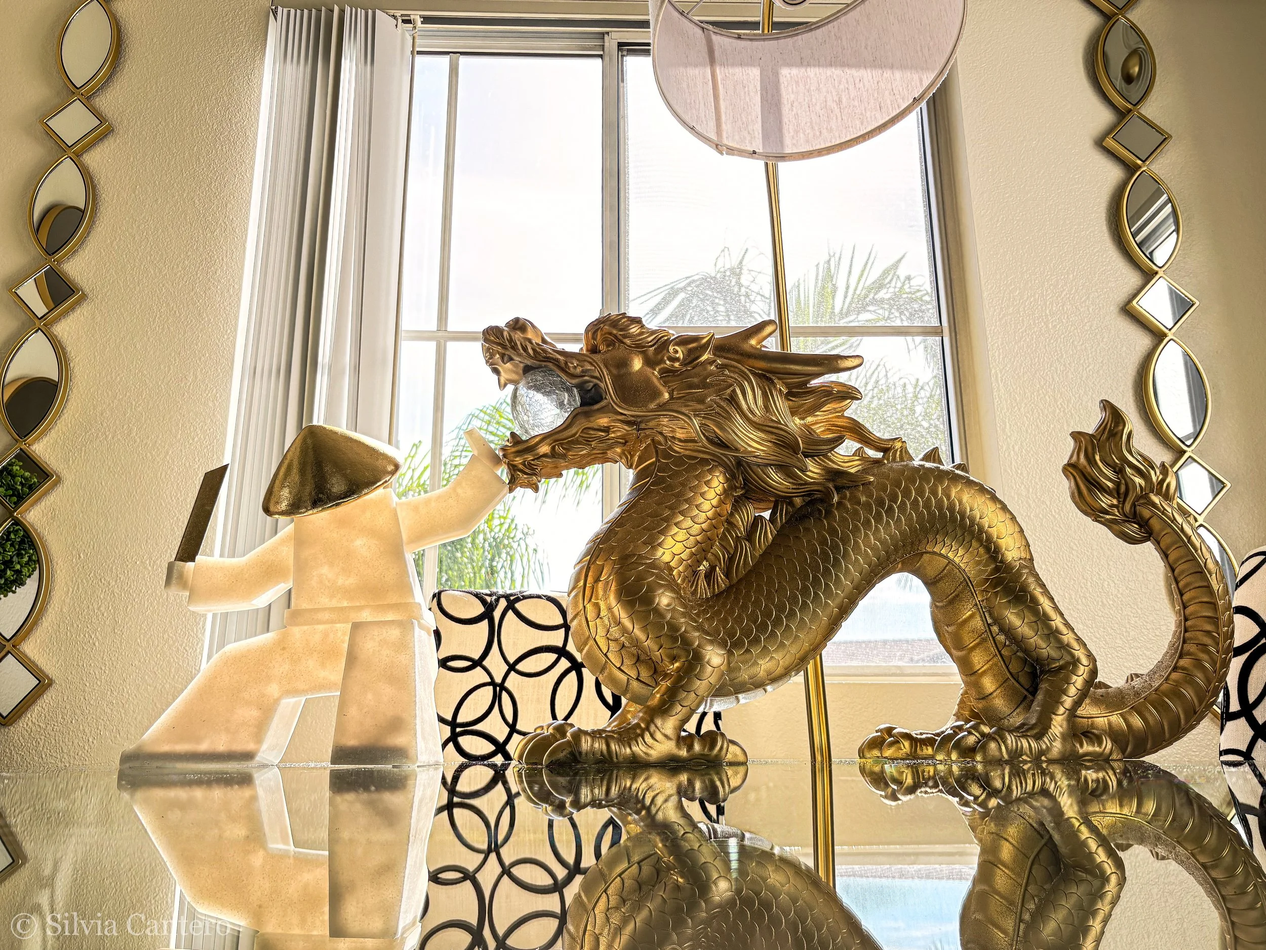 A decorative scene featuring a golden dragon statue, a white lamp, and a full-size mannequin torso, with large windows in the background letting in natural light and a cloudy sky beyond.