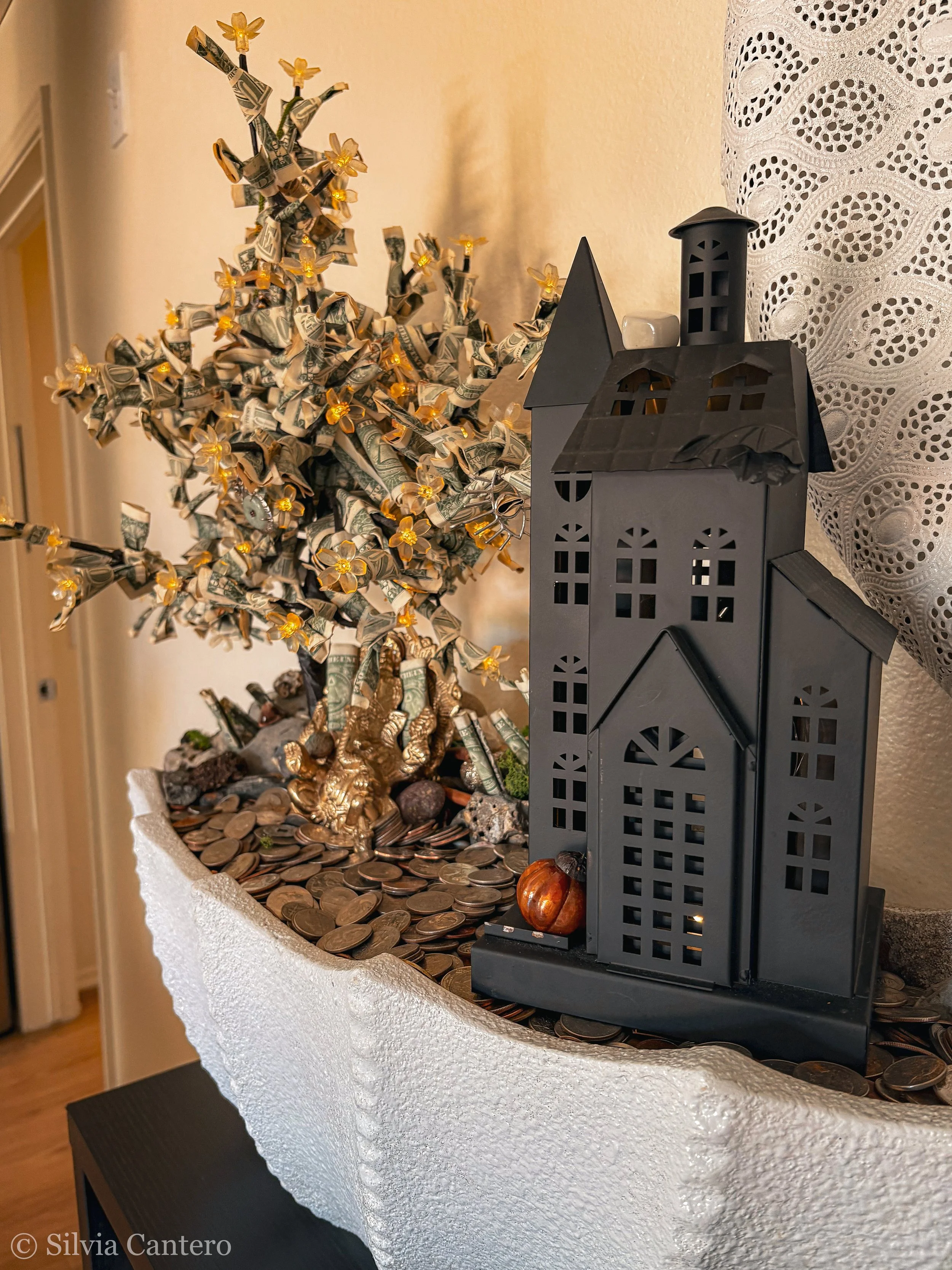 Decorative display with a black metal house-shaped lantern set on a white surface, surrounded by coins, a small pumpkin, and a metallic gold figurine of a bear. In the background, there is a money tree with rolled dollar bills and small flower lights