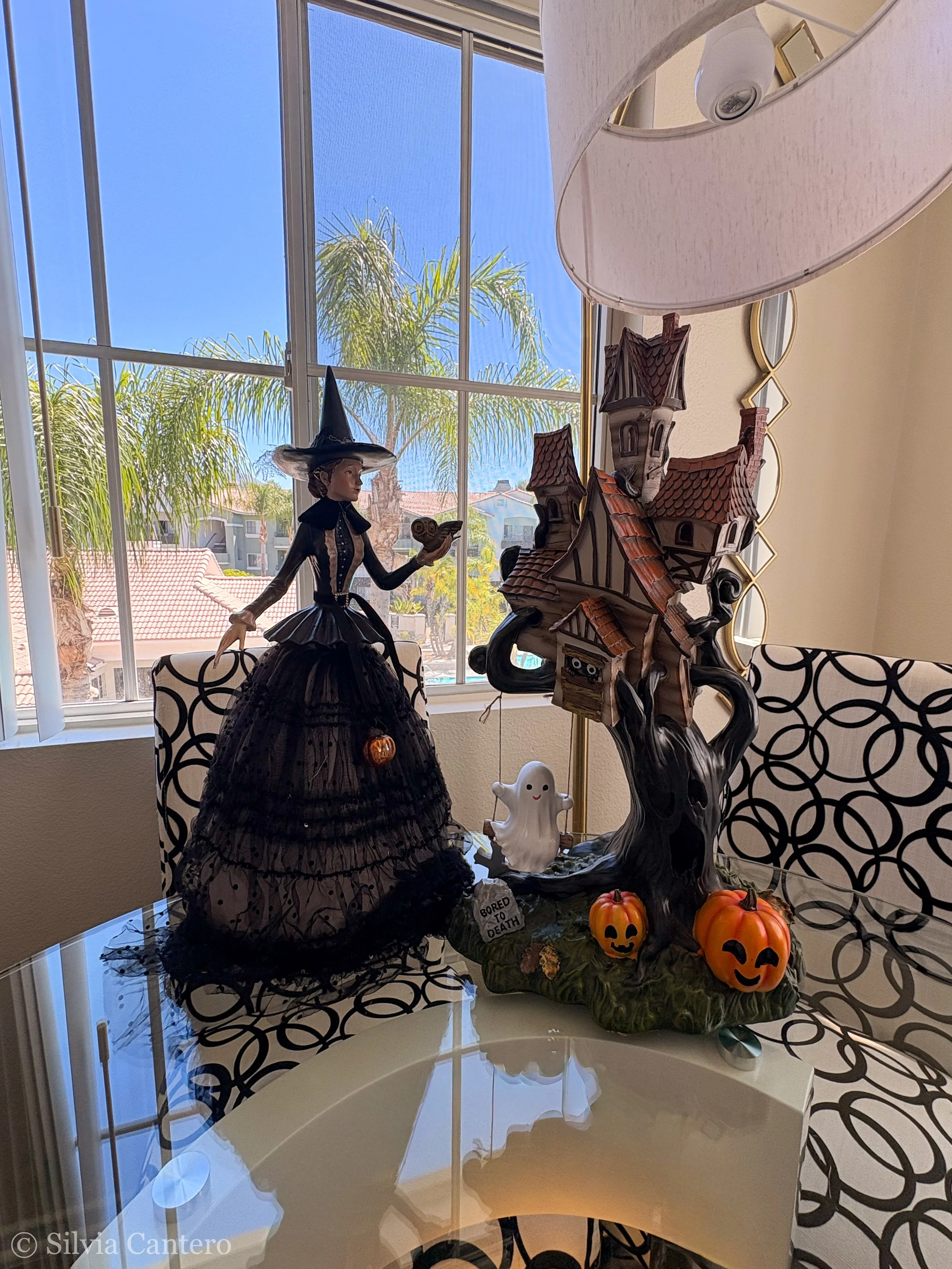 A Halloween decoration display on a table near a window, featuring a witch figurine wearing a black dress and hat, a haunted house sculpture, a smiling ghost figure, two small pumpkins with faces, a tree, and a sign that reads 'Bored to Death.' Insid