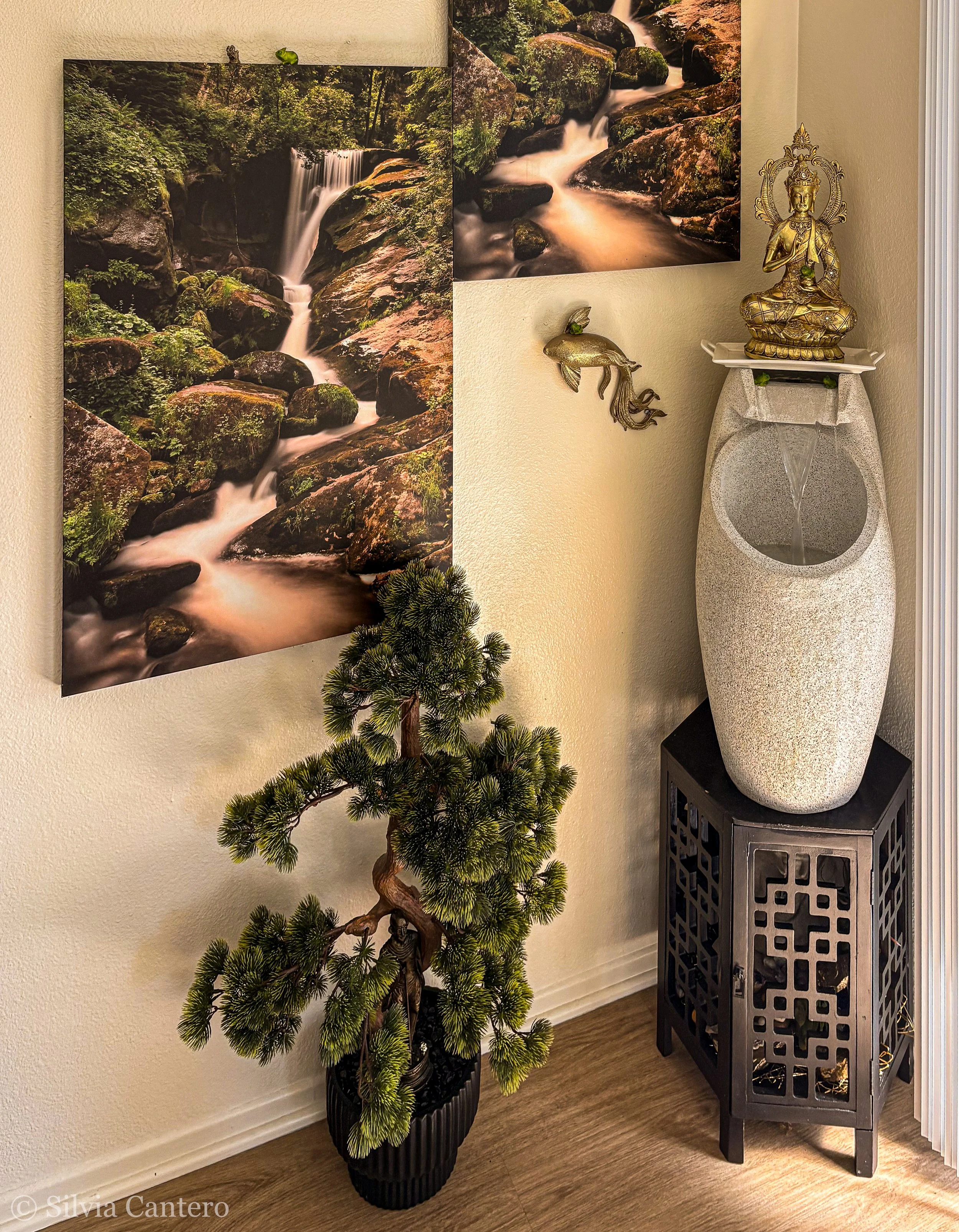 Interior decorative scene featuring a tall, potted pine tree, a three-panel landscape poster of a forest and waterfall, a brass statue of a deity or spiritual figure, and a large ceramic water feature with a carved black stand.
