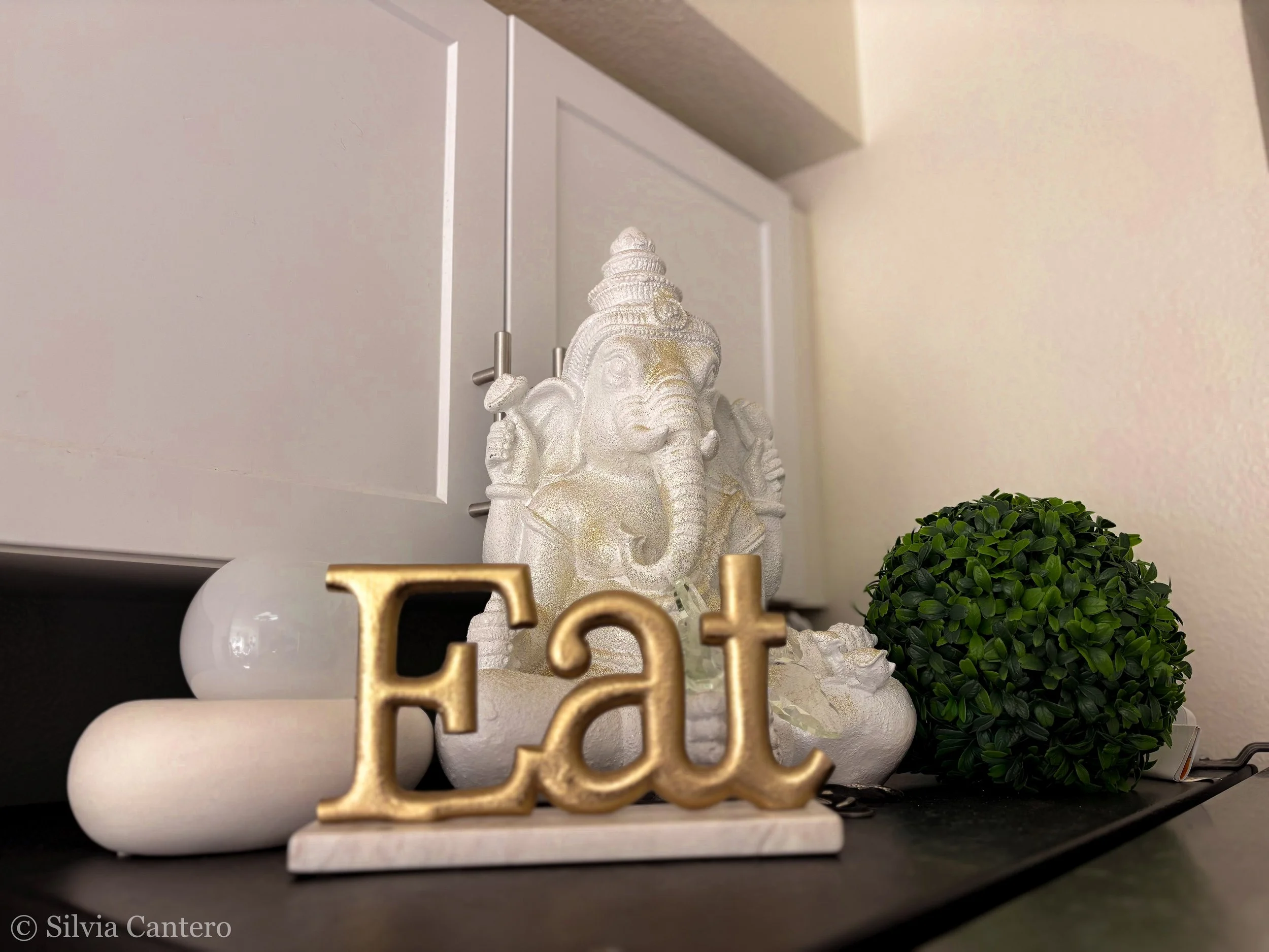Decorative display featuring a white Ganesha elephant head figurine with gold accents, a gold-colored 'LOVE' sign, a white candle, and a rounded green plant on a black surface in front of a white wall and cabinet.