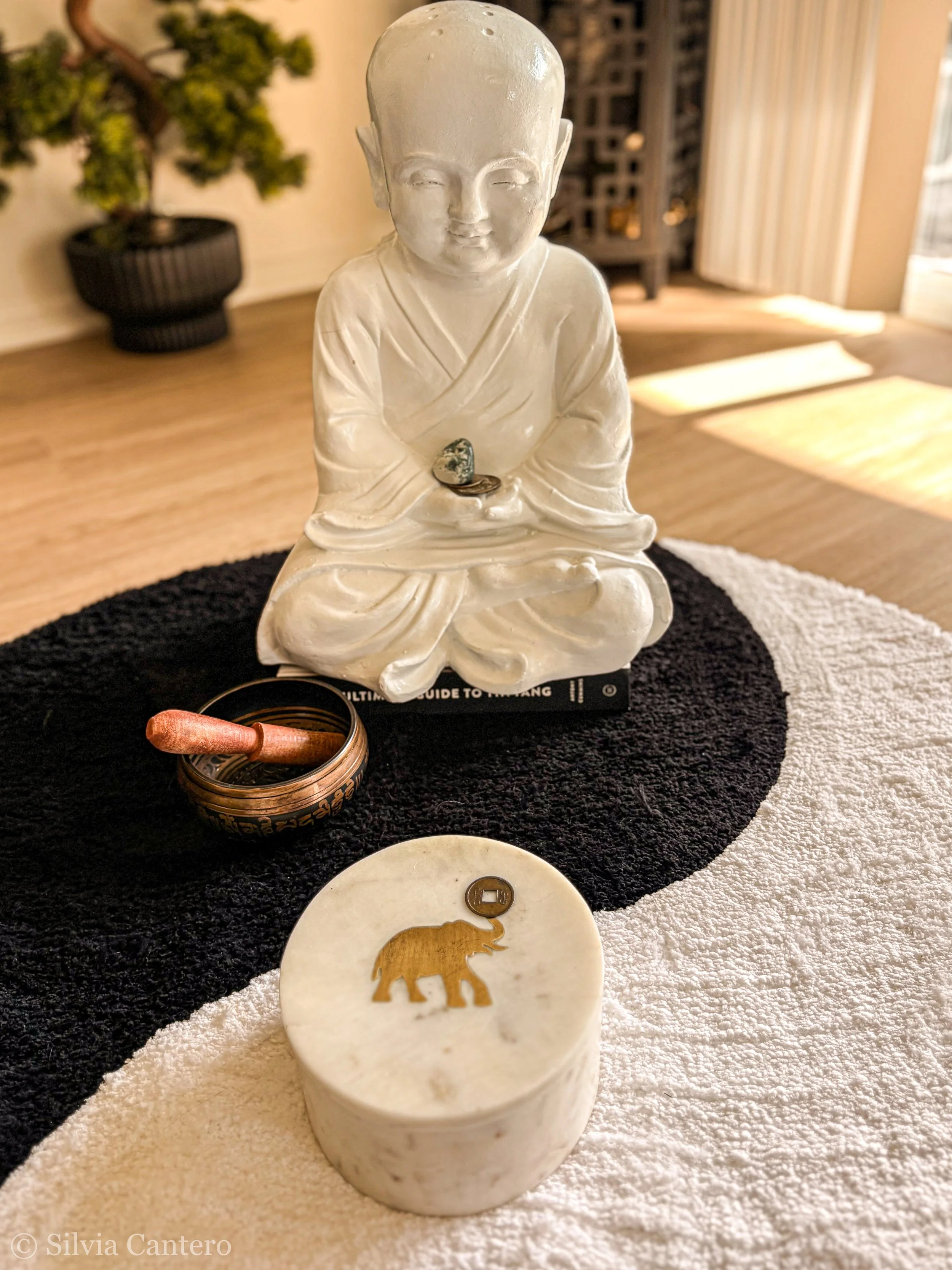 A white figurine of a seated person with a bald head, wearing traditional clothing, and holding small stones, placed on a black and white circular cloth with other small objects including a carved dish with a gold elephant and a coin.