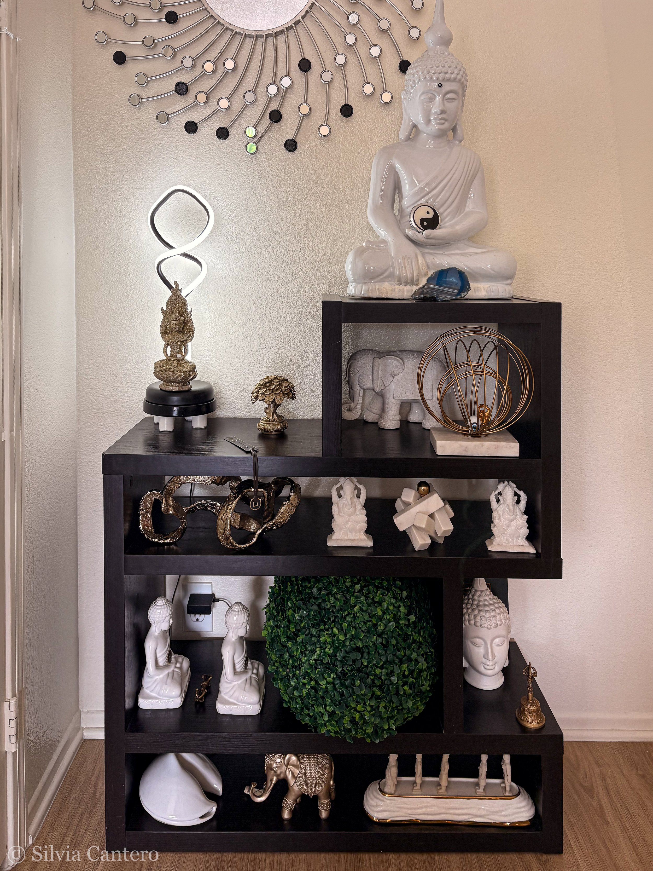 Decorative black wooden shelf with various Asian-inspired ornaments, sculptures, and decor including Buddha figures, a yin-yang symbol, a tree sculpture, a white helmet, a gold elephant, and a white dish, with a green plant and wall art in the backgr