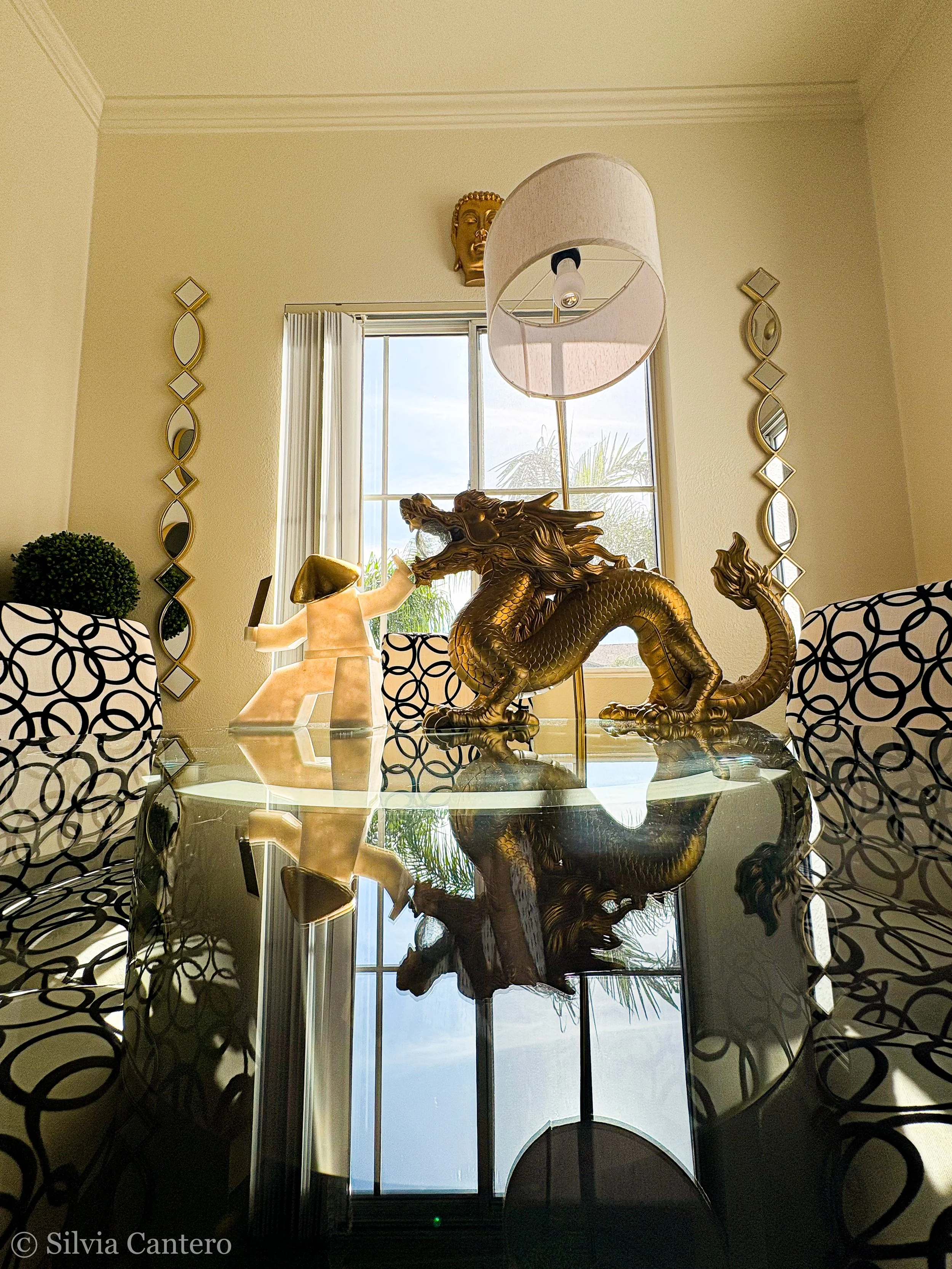A decorative living room scene with a golden dragon sculpture on a glass table, a beige and white lamp, and gold framed mirrors on a cream-colored wall.