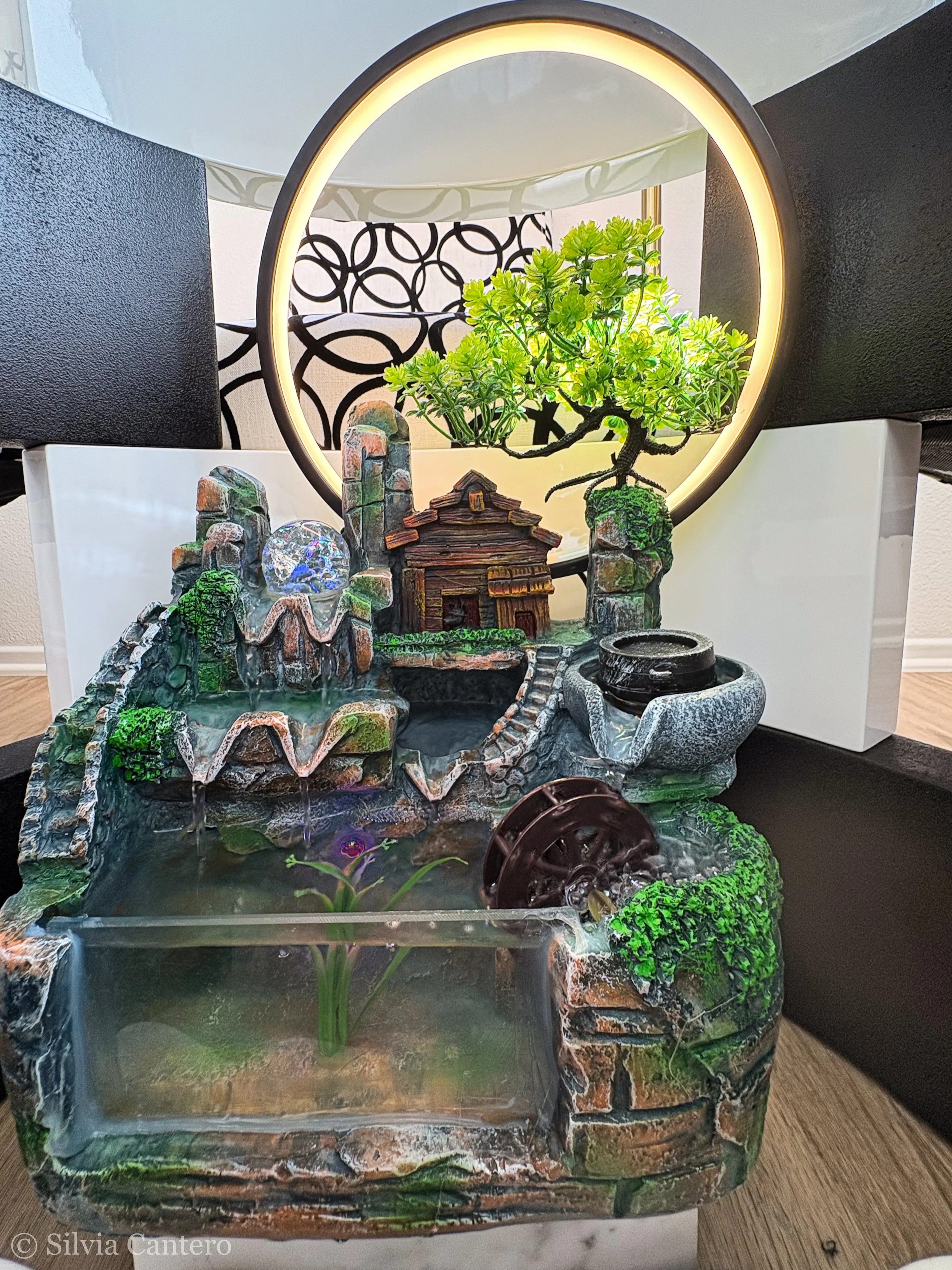 Decorative tabletop fountain with miniature house, rocks, and a small waterfall, featuring a circular LED light, placed on a black surface with cushions and patterned wall in background.