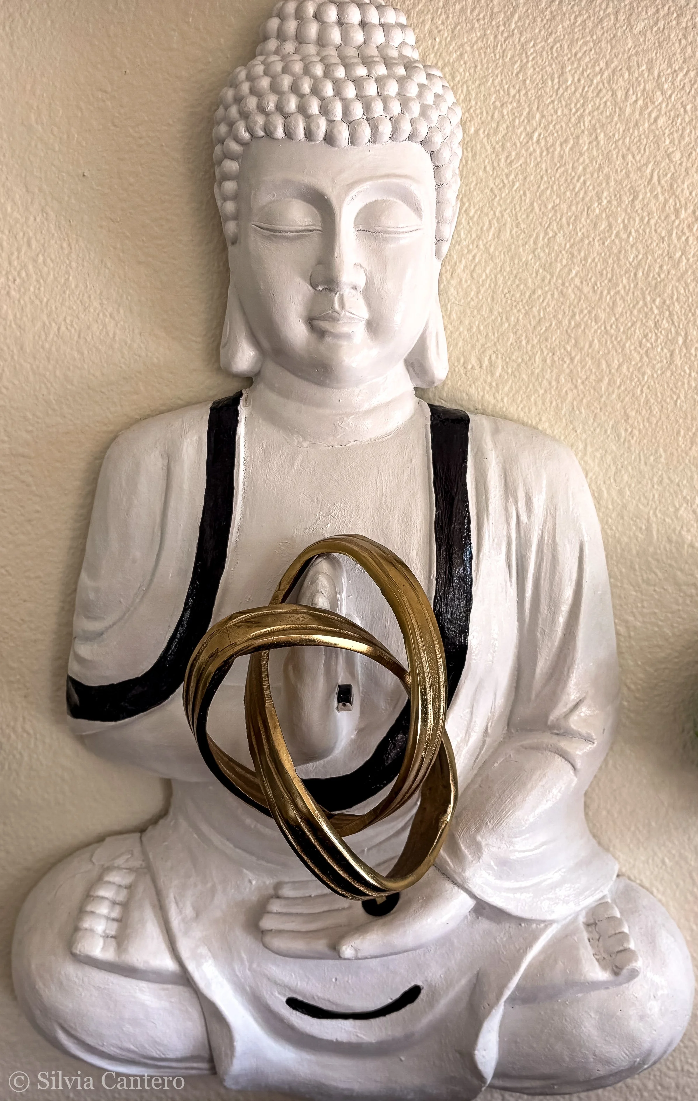 White Buddha statue with closed eyes, black painted accents on the robes, and intertwined gold rings hanging from the chest, situated against a beige wall.