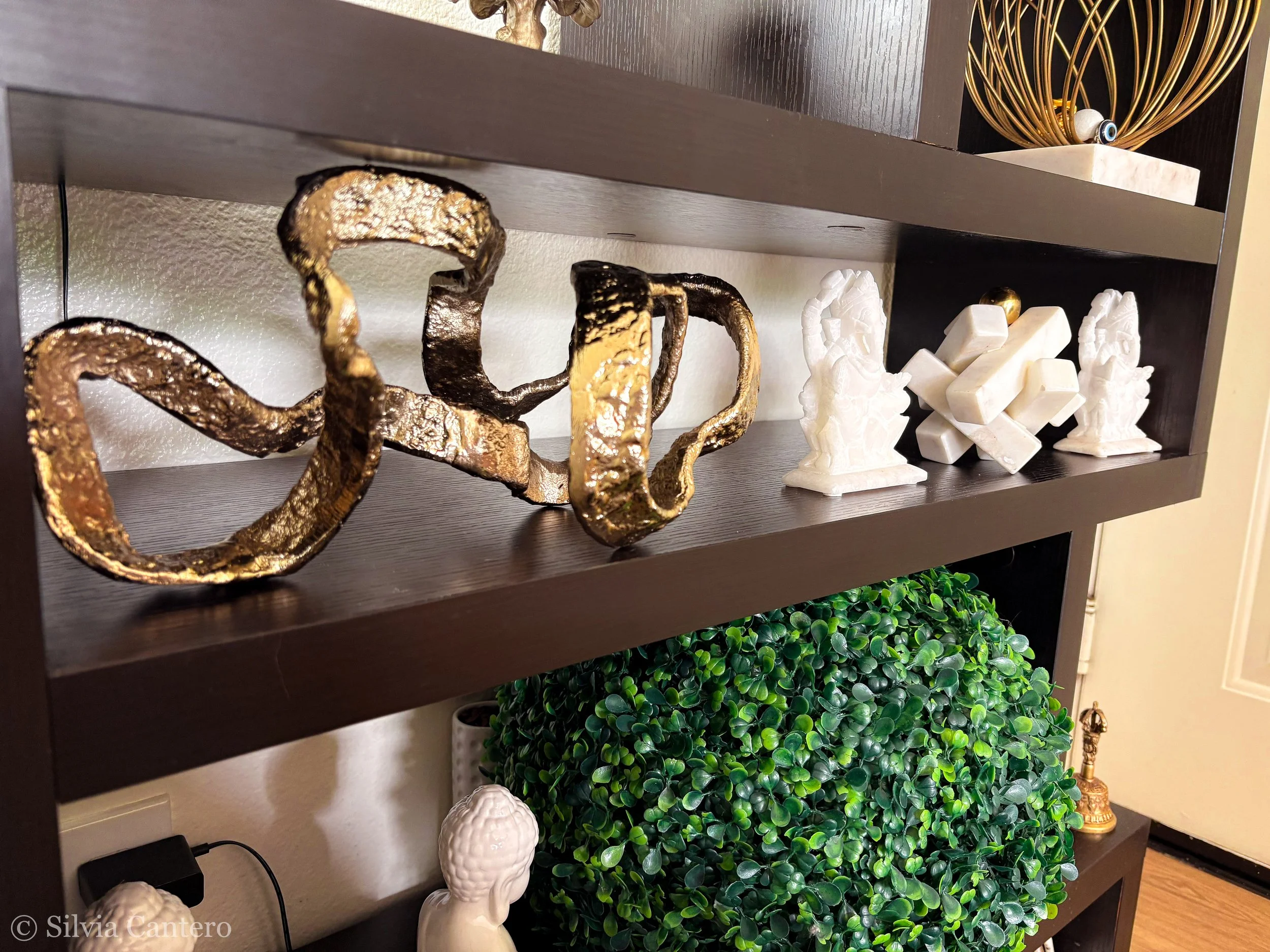 Decorative items on black shelves including golden metallic sculpture, white Buddha statues, and a glass bowl with a white marble base, with a faux green plant underneath.