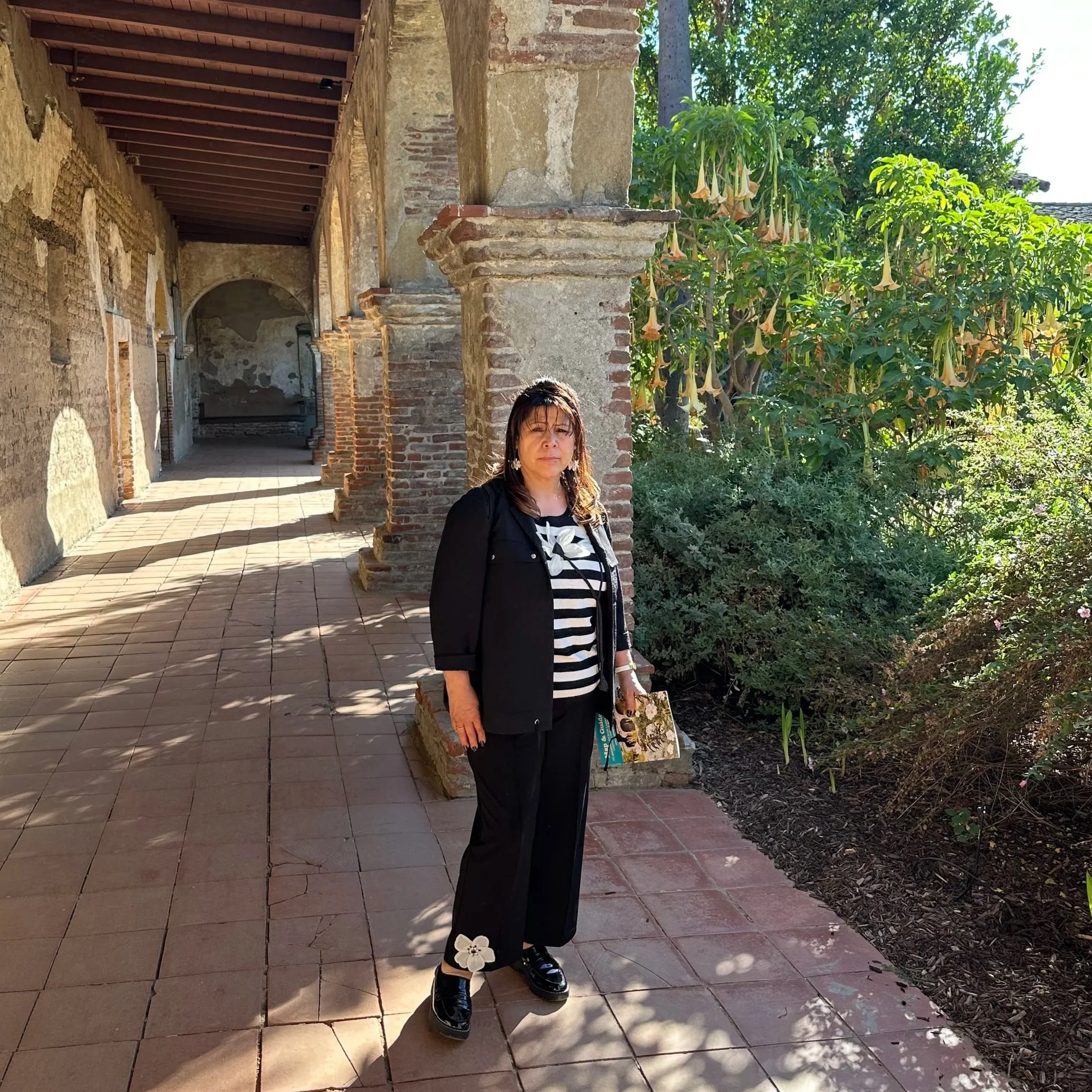 A woman standing outdoors beside a stone column and lush green plants, wearing a black jacket and striped shirt with black shiny shoes, holding a magazine or booklet.