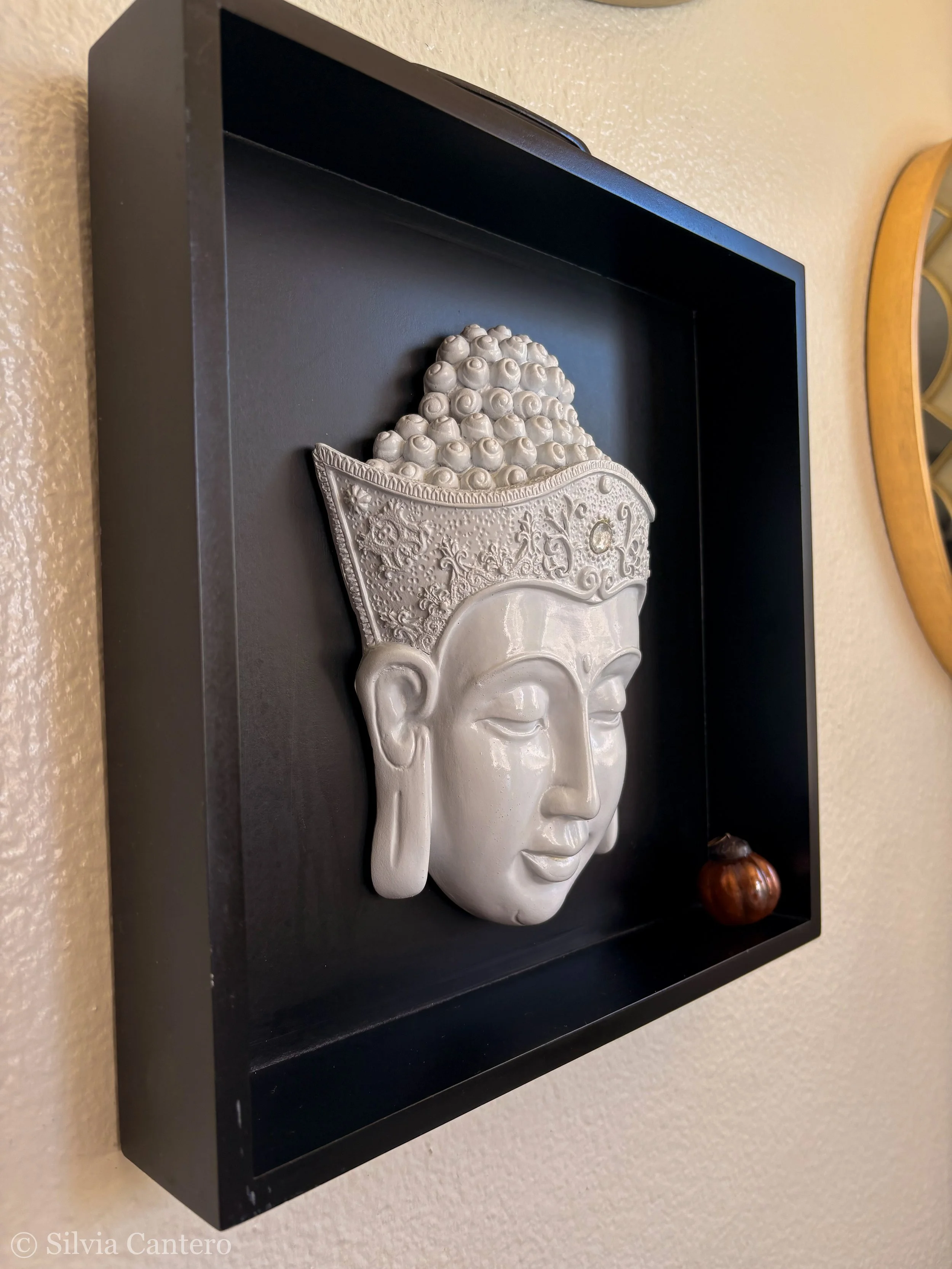 A decorative wall art piece featuring a white 3D mask of a face with closed eyes, wearing an ornate crown or headdress, mounted on a black shadow box frame, with a small round object on the right side of the frame.