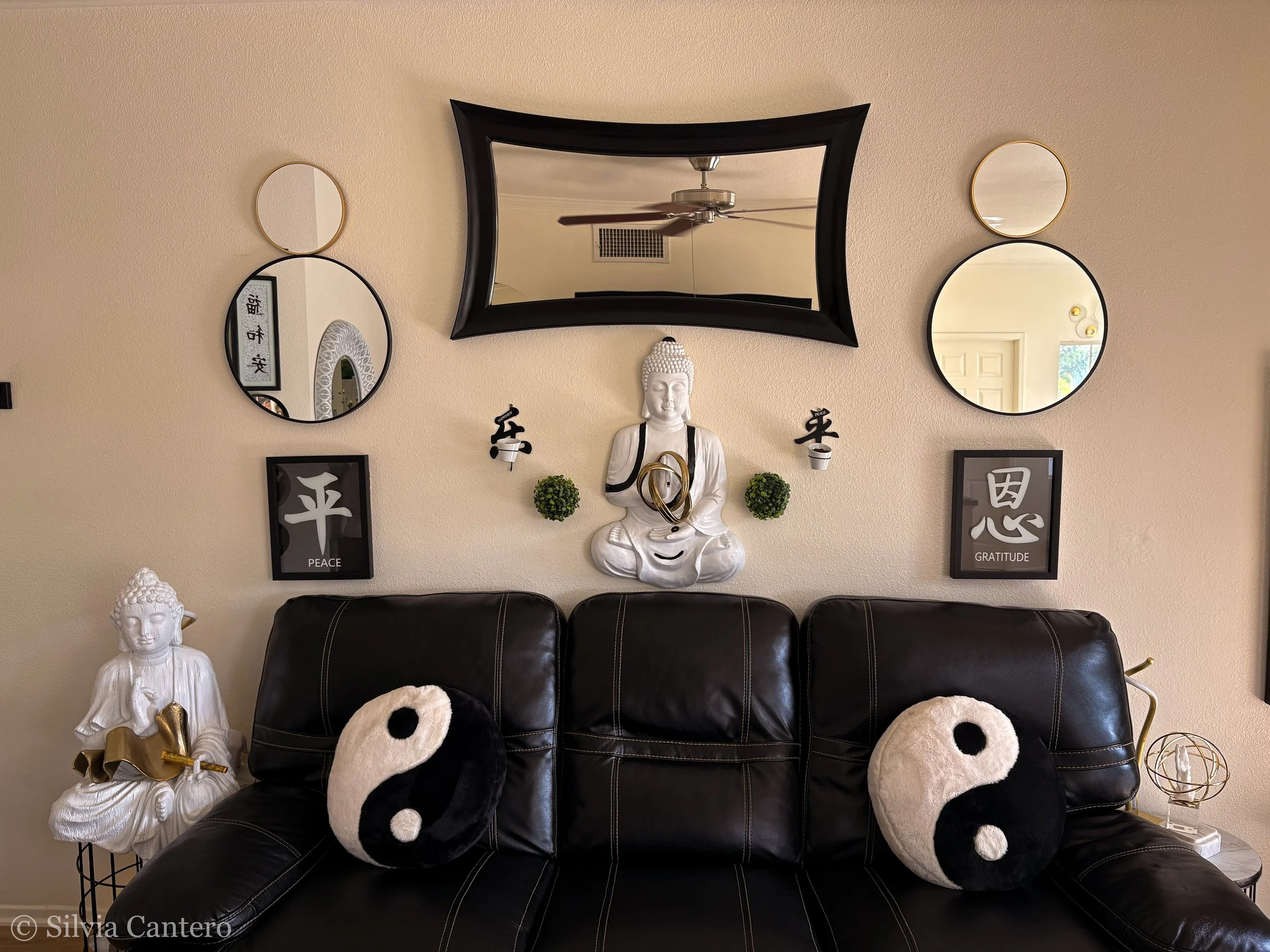 Living room wall decor featuring three mirrors, a white Buddha statue, framed artwork with Chinese characters labeled "Peace" and "Gratitude," two small green plants, plush panda cushions on a black leather couch, and a ceiling fan reflection in the 