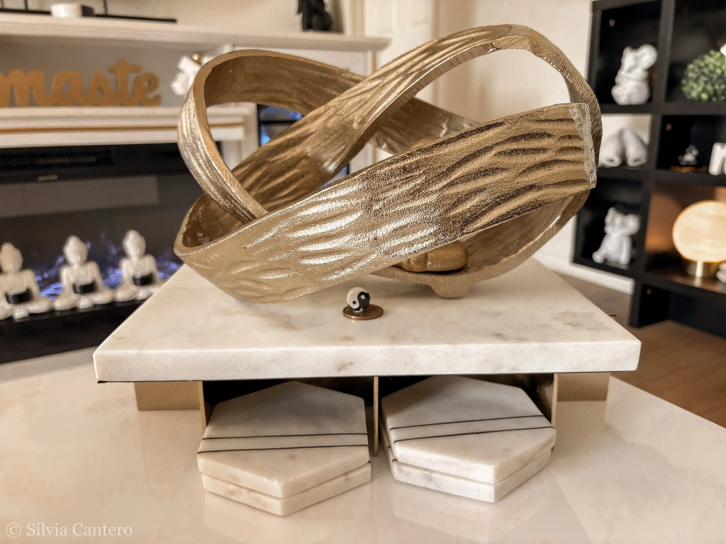 A decorative wooden sculpture with flowing, intertwined shapes sits on a stand above a white marble base. In the background, a black shelf holds small figurines, decorative objects, and a sign spelling "matte" in gold letters.