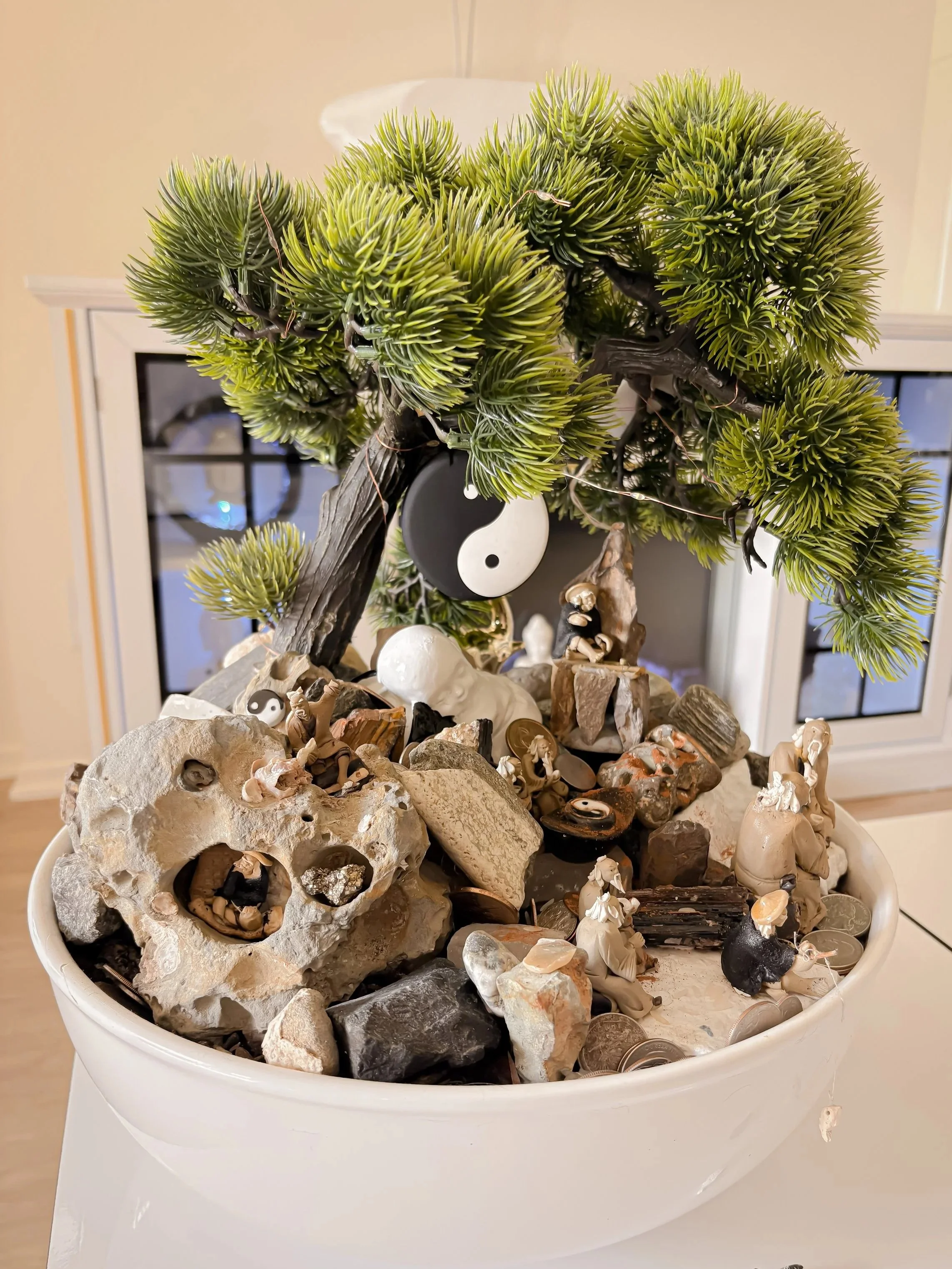 A white planter filled with various rocks, shells, and small figurines, topped with a small artificial tree with green needle-like leaves.