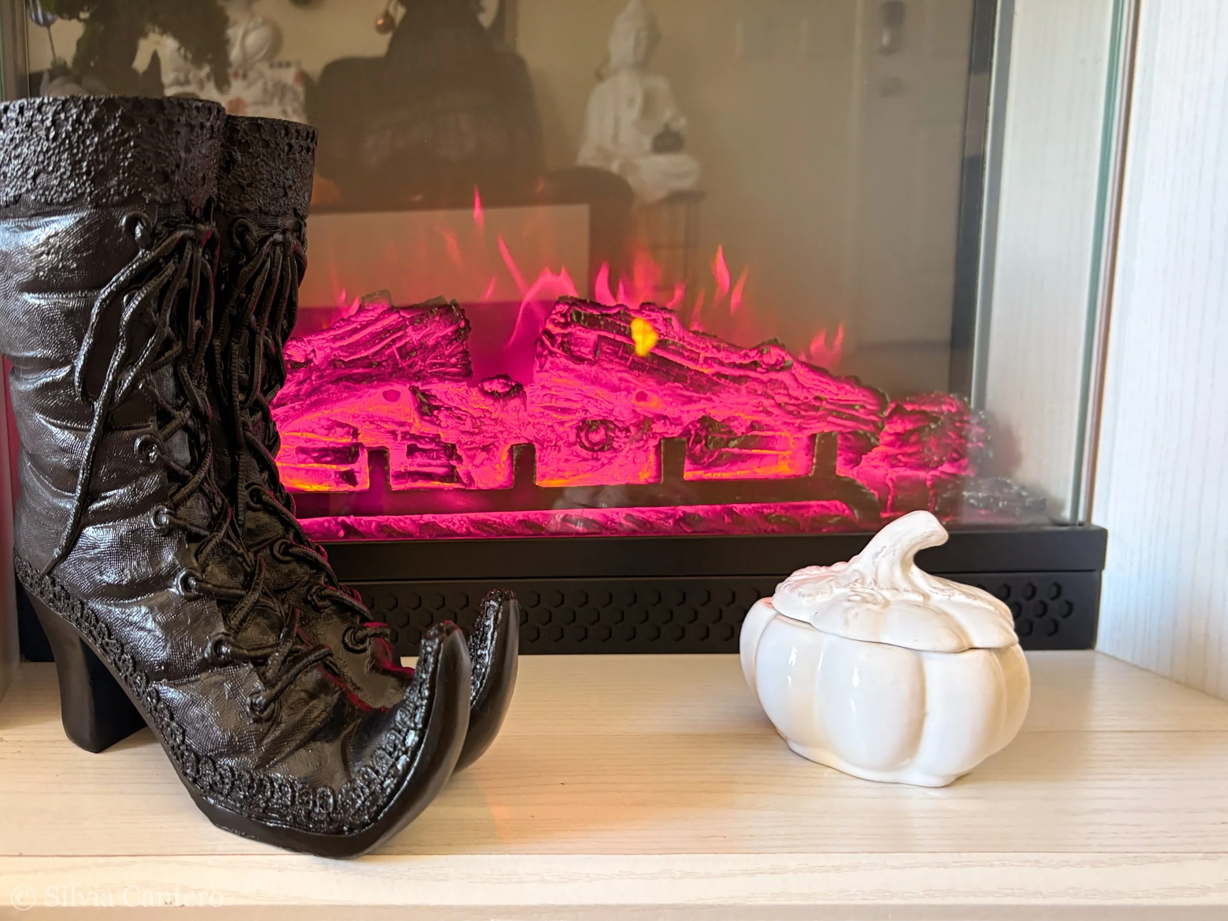 Black leather lace-up boot placed on a light-colored table, with a white ceramic container shaped like a pumpkin beside it, in front of a glass-enclosed fireplace with pink and red glowing artificial embers.