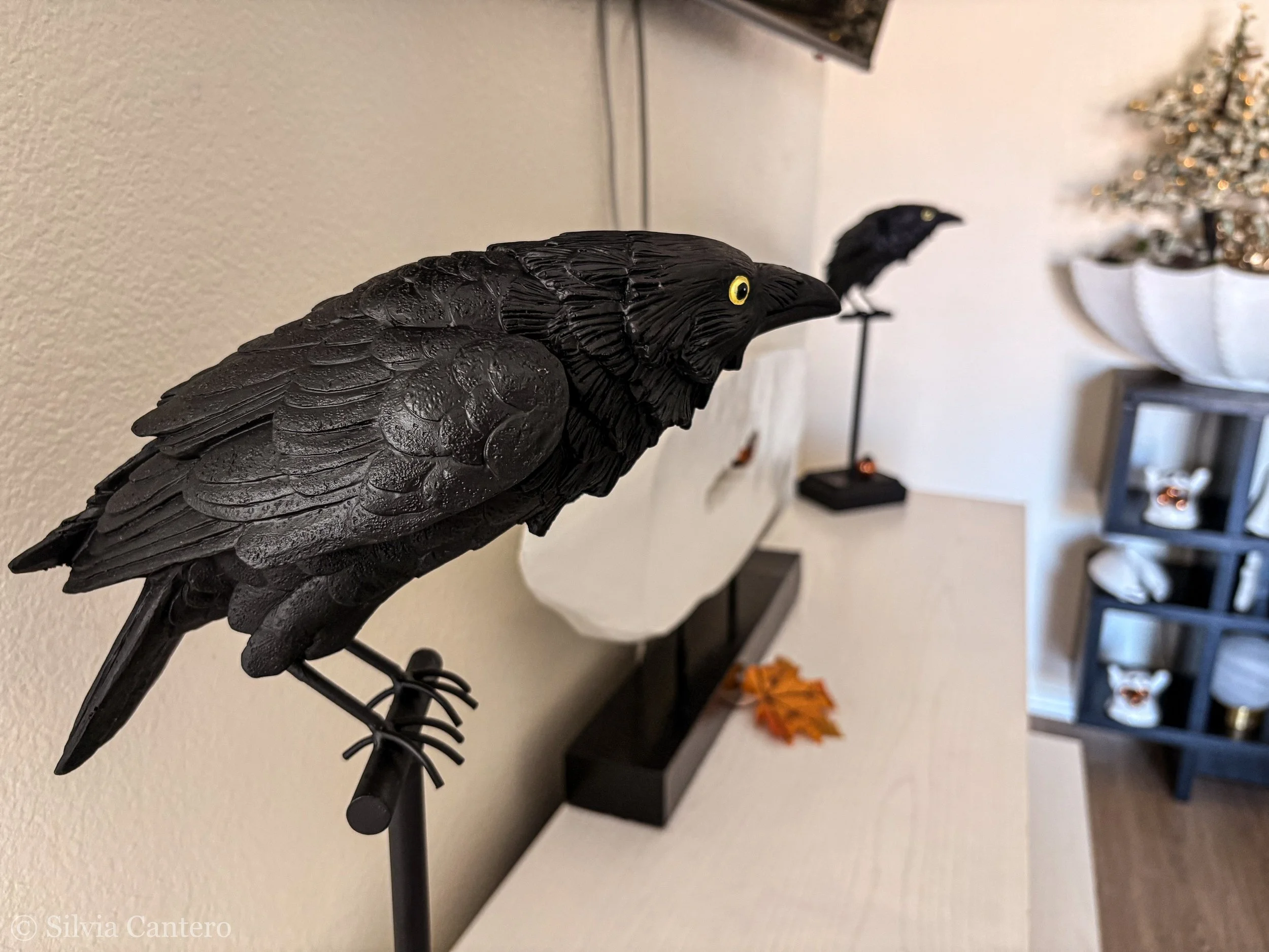 A black three-dimensional bird sculpture with detailed feathers and bright yellow eyes, mounted on a stand, with a blurred background including a Christmas tree and other decorative items.
