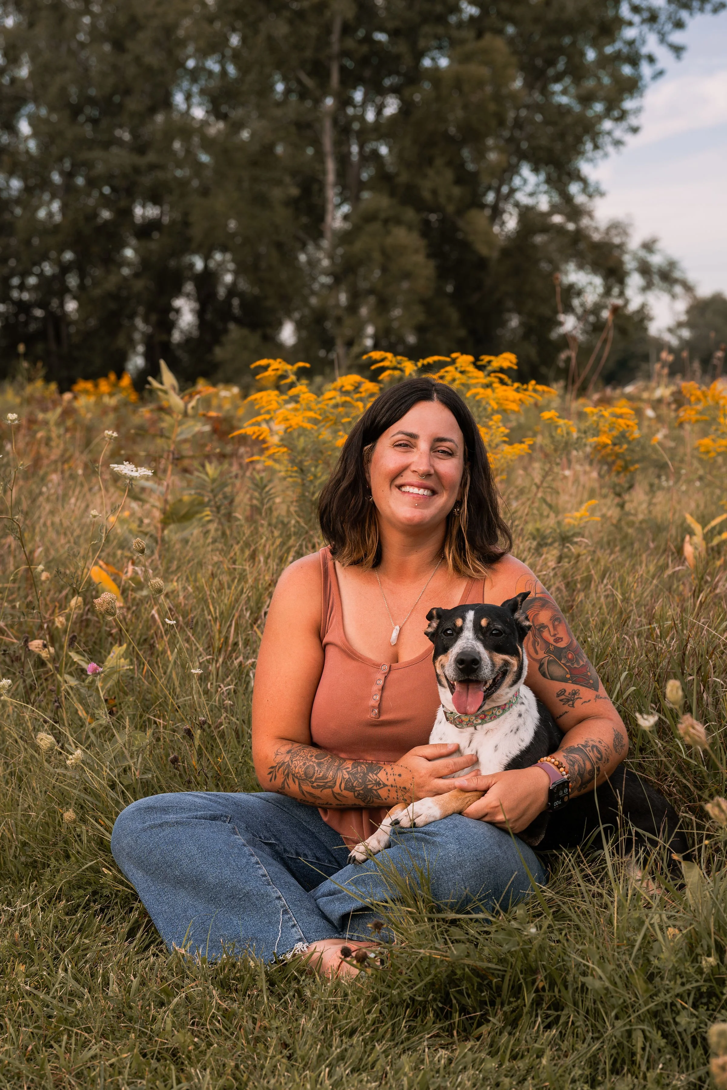 A woman with tattoos and dark hair sitting cross-legged in a field of wildflowers, smiling, and holding a black and white dog with a colorful collar. Trees and a cloudy sky are in the background.
