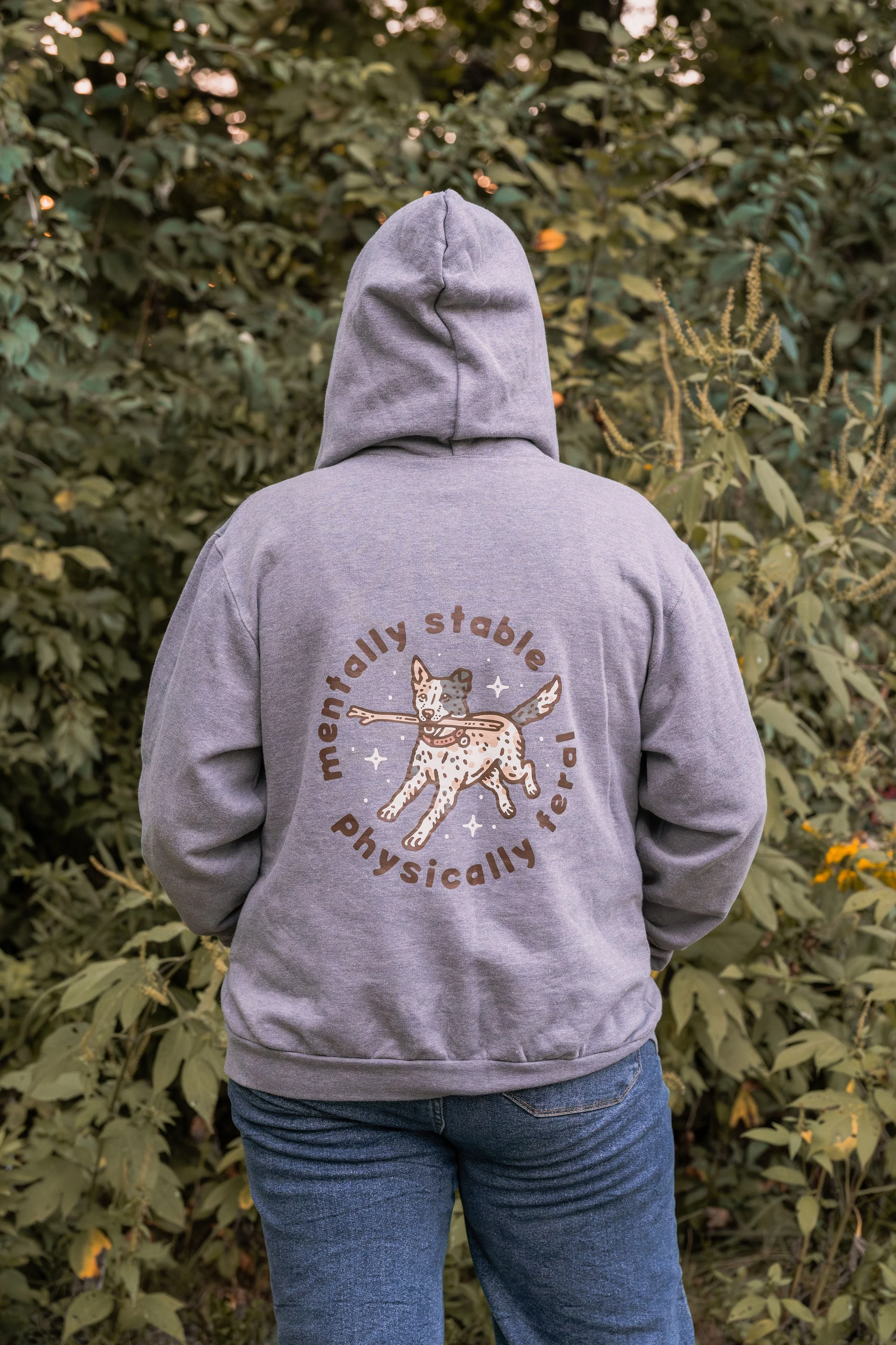 Person wearing a gray hoodie and blue jeans standing with back to camera in front of green leafy bushes. The hoodie has a circular logo featuring a dog and the words 'mentally stable, physically fit'.